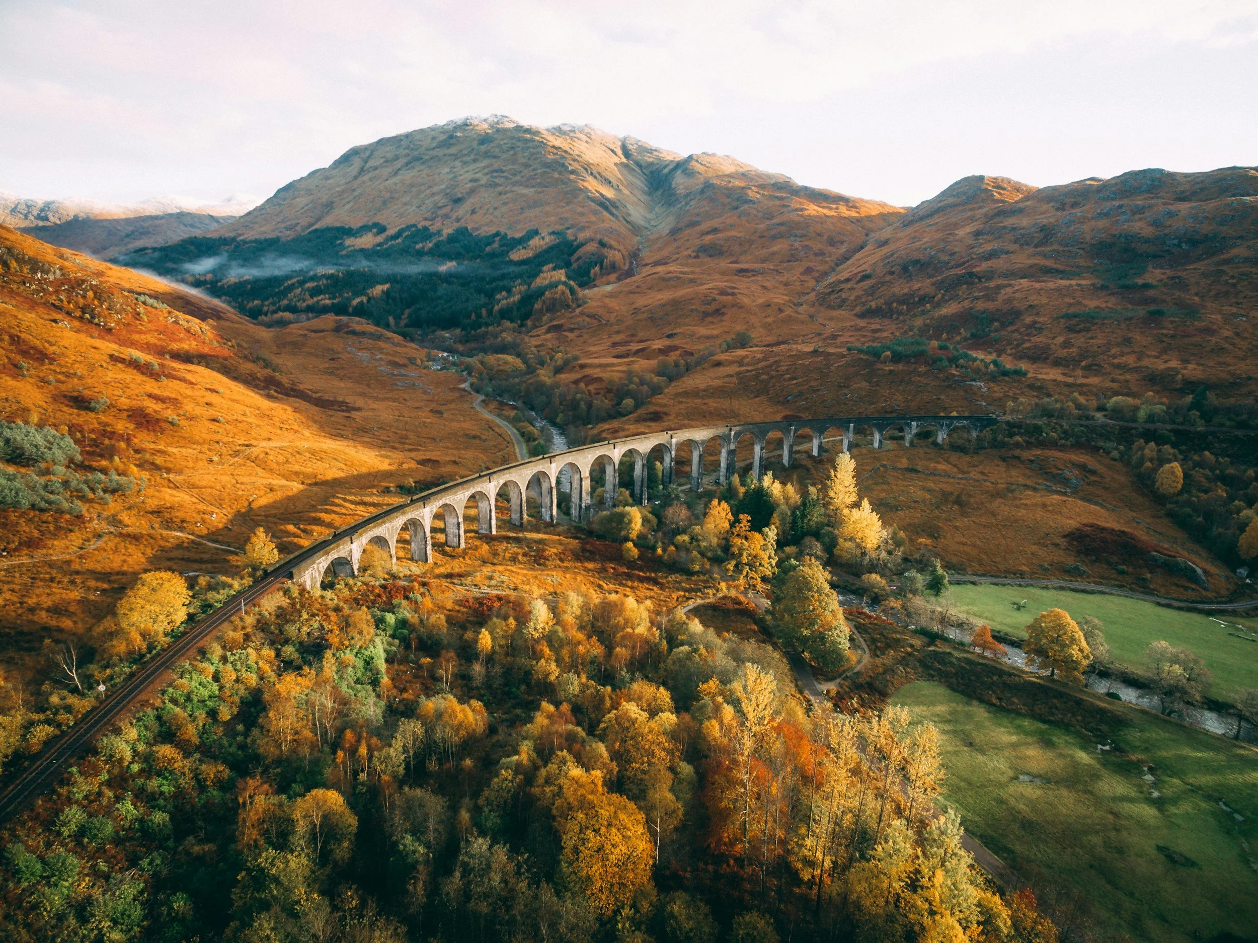 A scenic railway viaduct crossing a valley surrounded by rolling hills covered in autumn-colored trees and foliage, with mountains in the background under a partly cloudy sky.