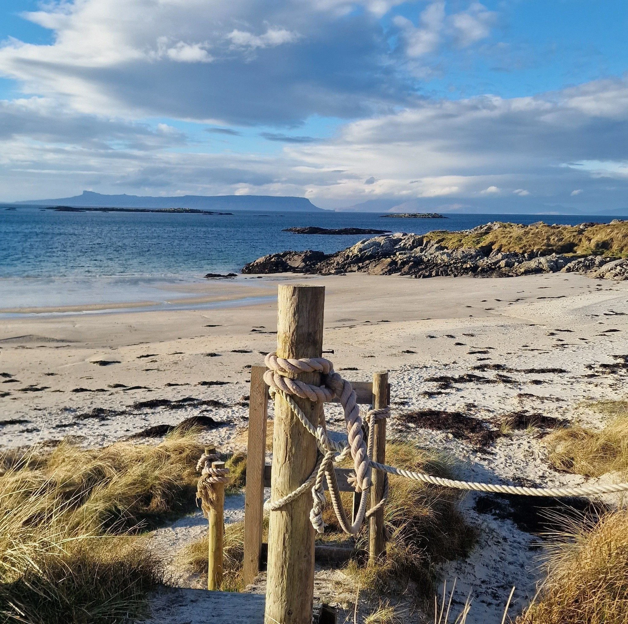 A view of a sandy beach with grassy dunes and rocks, a rope fence, calm ocean water, distant islands, and a partly cloudy sky.