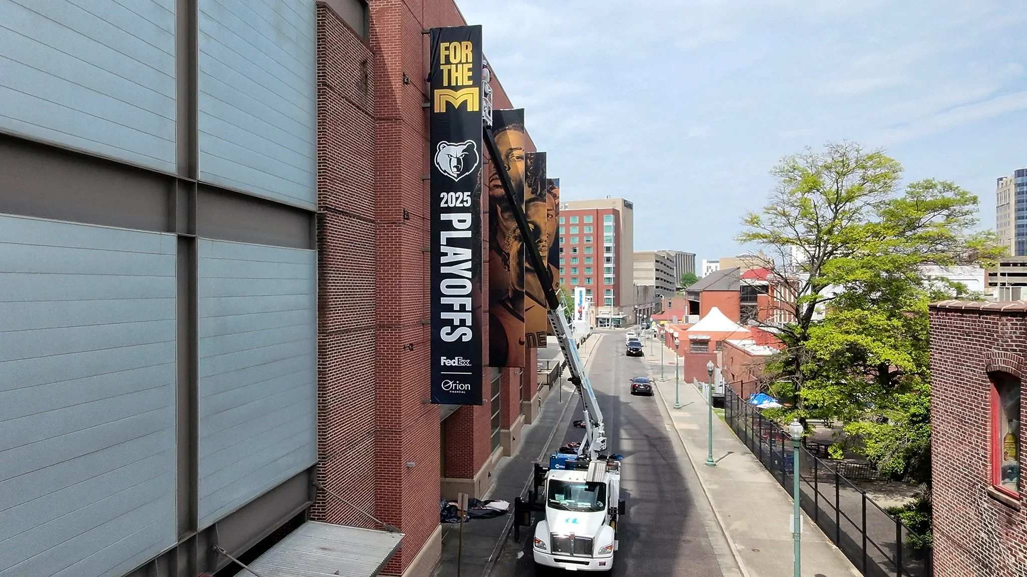 A city street scene with a large vertical banner on the side of a brick building promoting the 2025 Memphis basketball playoffs, sponsored by FedEx and Orion Financial, with a crane lifting a worker near the banner and several trees, cars, and buildi