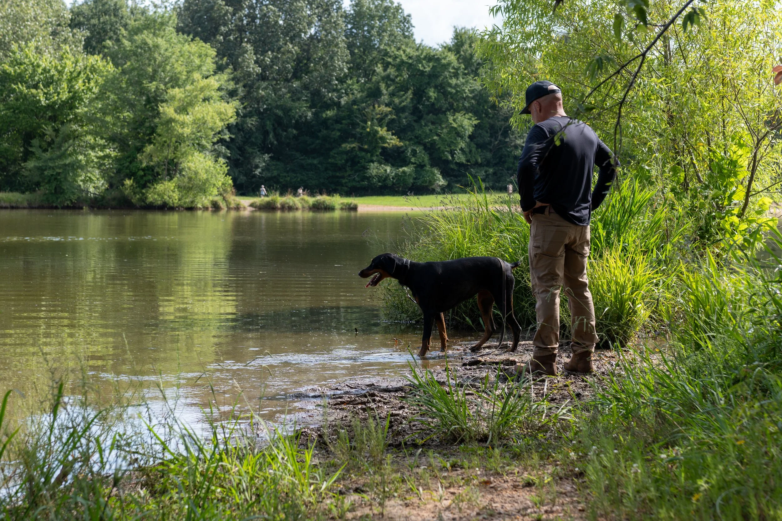 A man in a black cap, black long-sleeve shirt, and beige pants stands on a riverbank with a black and tan dog, near green trees and grass, overlooking the water on a sunny day.