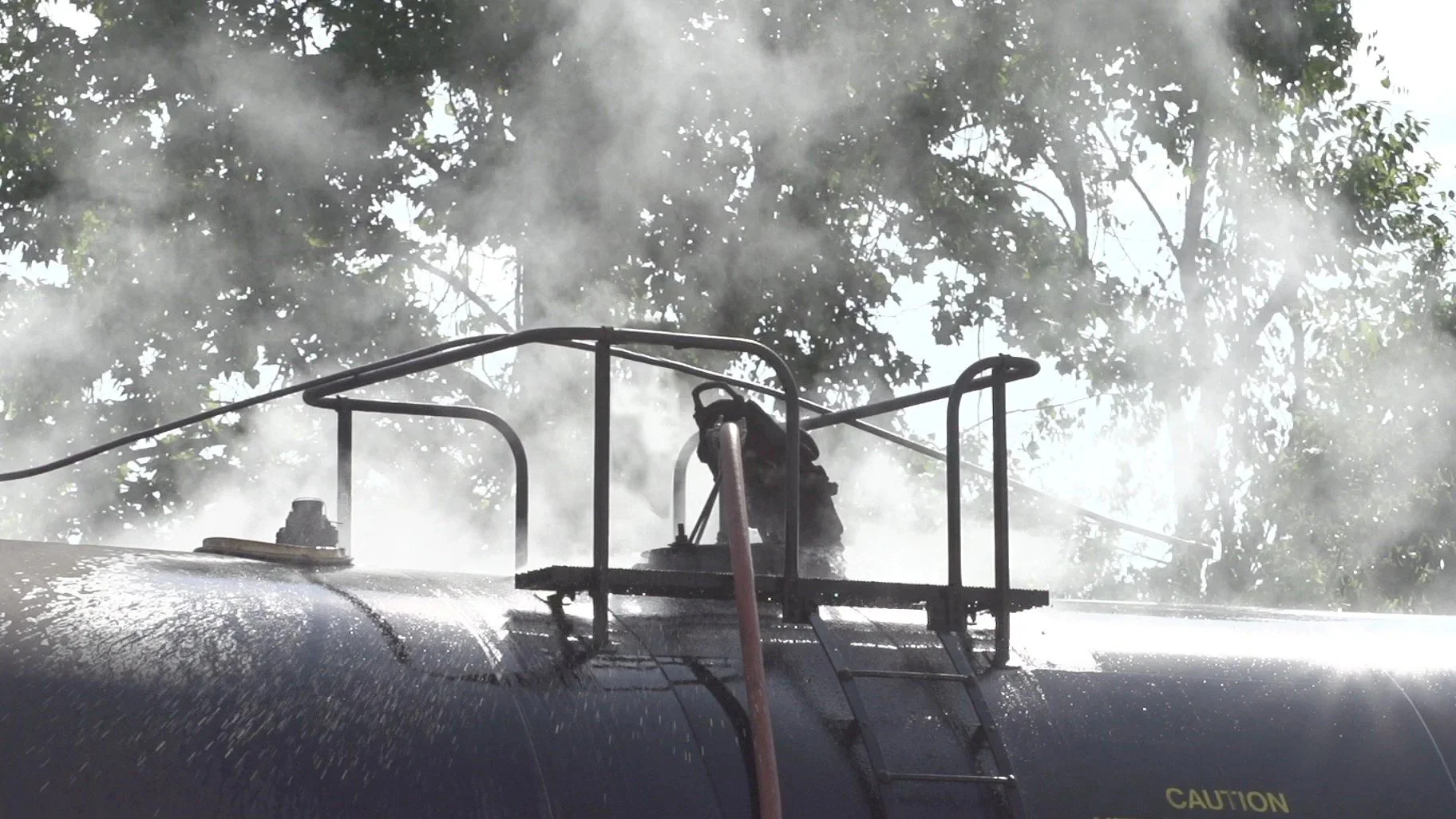 A firefighter wearing protective gear and helmet is on top of a water tanker, spraying water into the air, with steam and smoke rising and trees in the background.