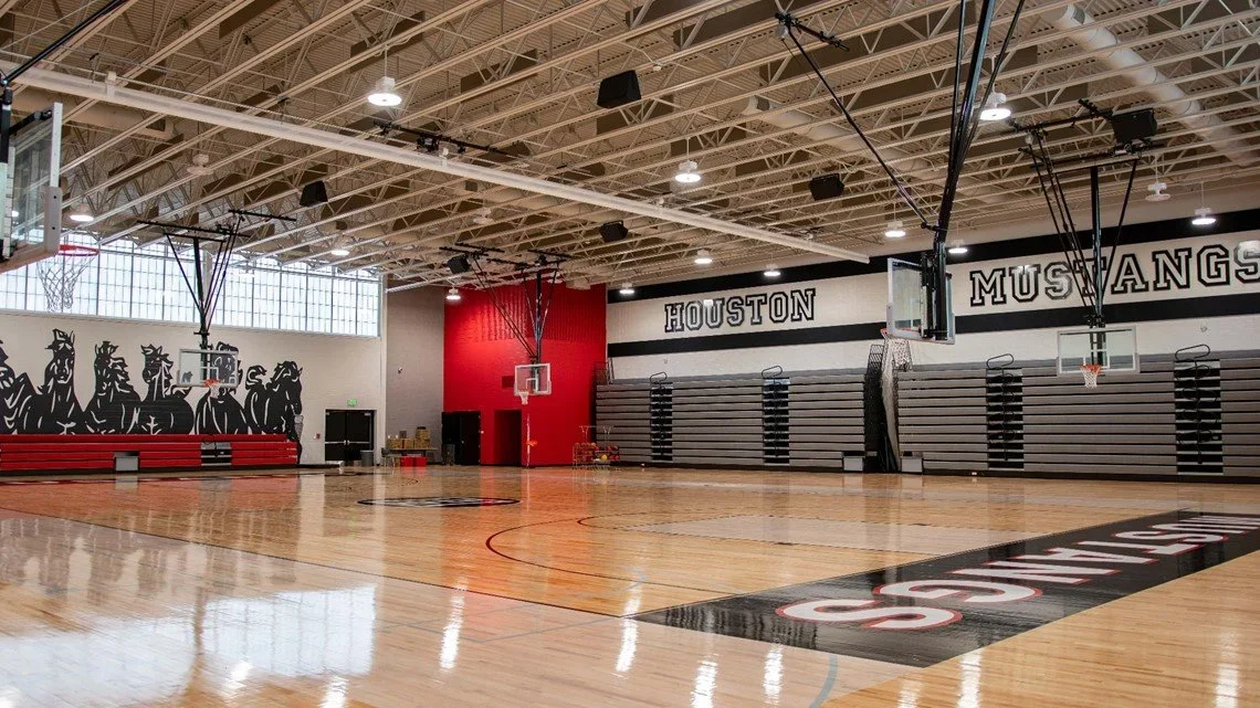 Empty basketball gym with wooden floor, two basketball hoops, gray bleachers, and wall decor reading "HOUSTON MUSTANGS".