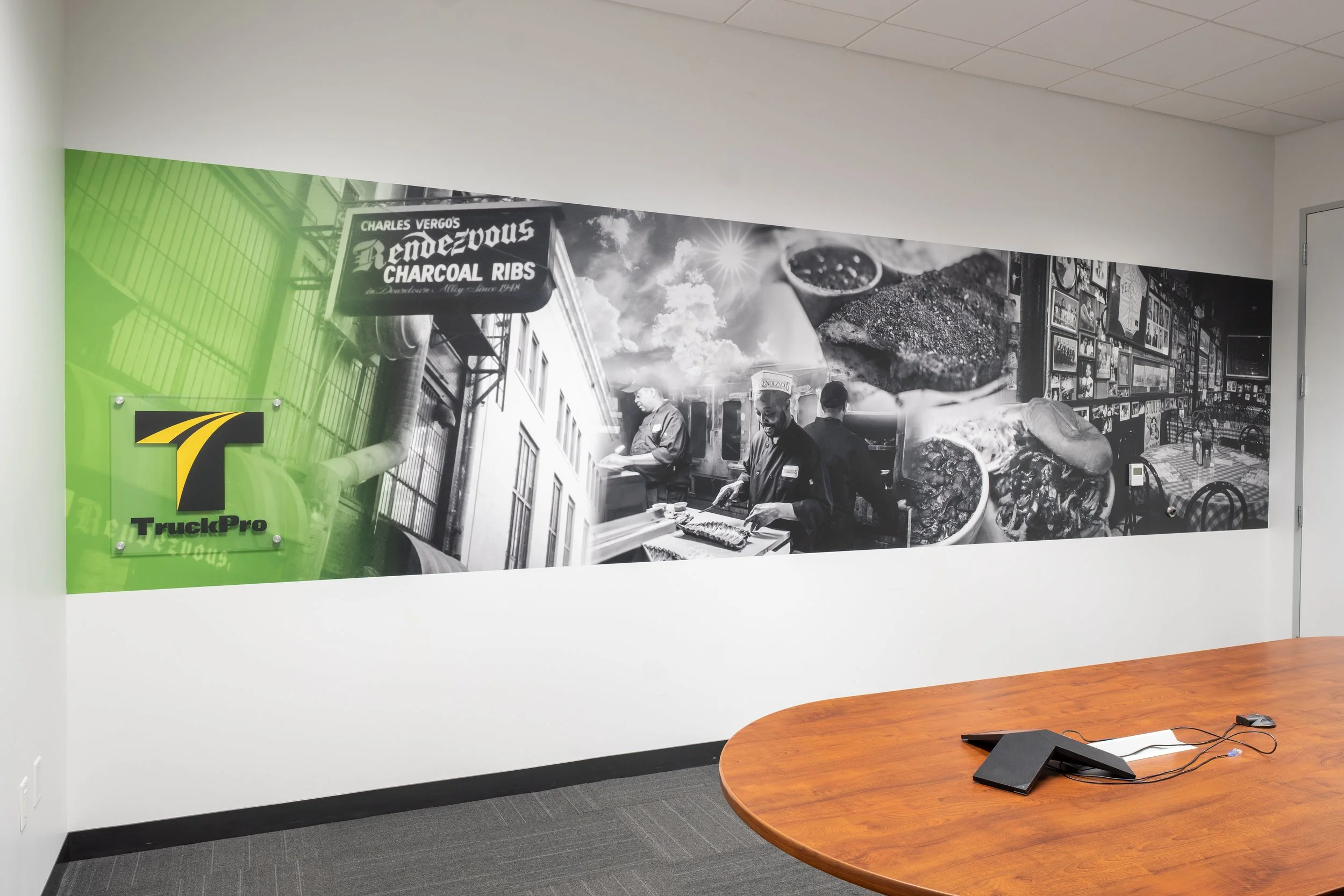 A large black and white mural on a white wall depicts a street scene with a sign reading 'Charles Vergo's Rendezvous Charcoal Ribs,' a chef preparing food, and various food images including ribs and a hot dog. A green logo with the letter 'T' and the