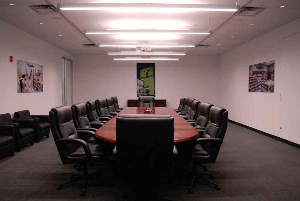 Empty conference room with a large oval wooden table surrounded by black executive chairs, with artwork on the white walls and ceiling lights.