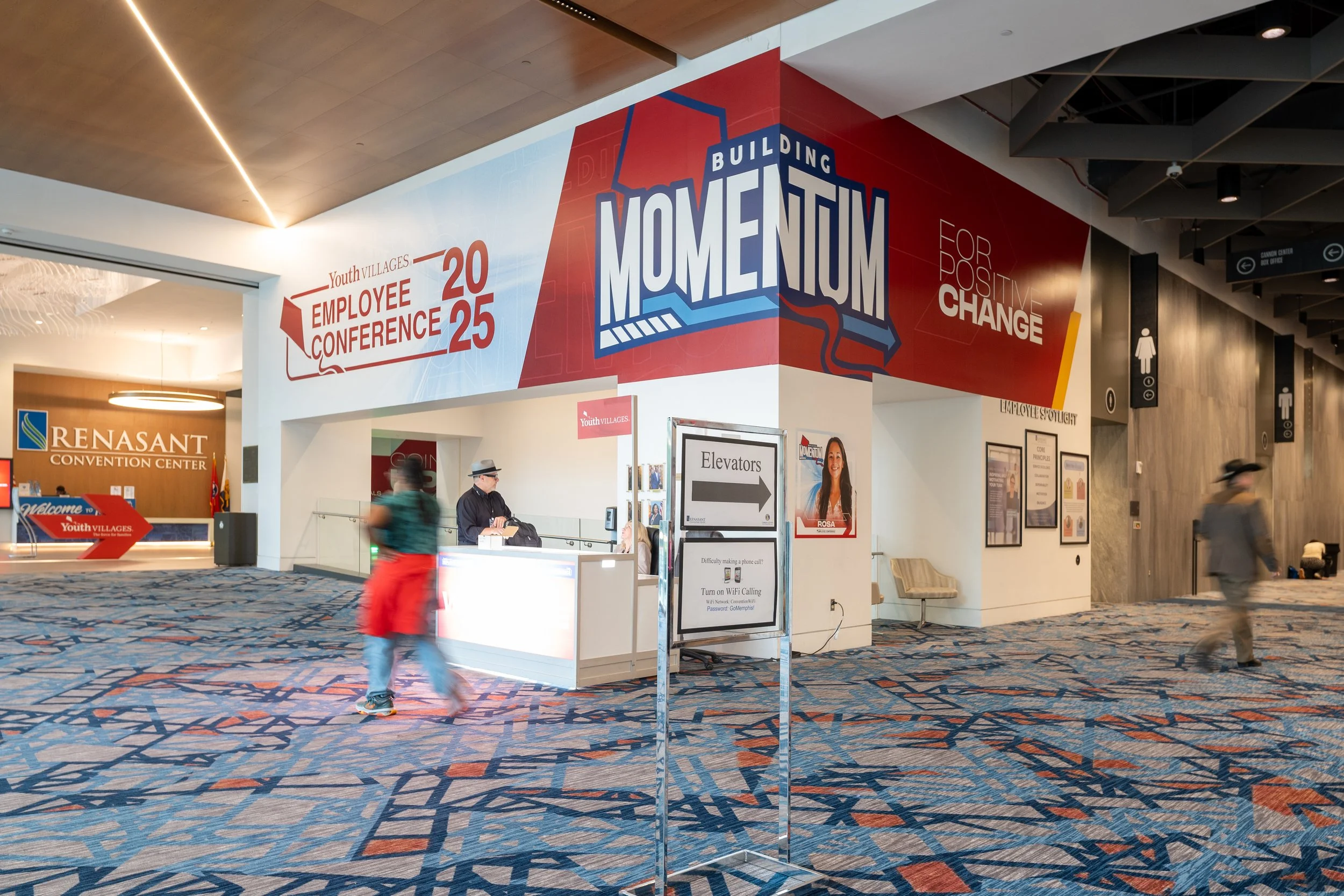 Interior of a convention center with signs for Youth Villages Employee Conference 2025, building momentum, and positive change. People are walking and waiting at the reception area, with a colorful patterned carpet on the floor.