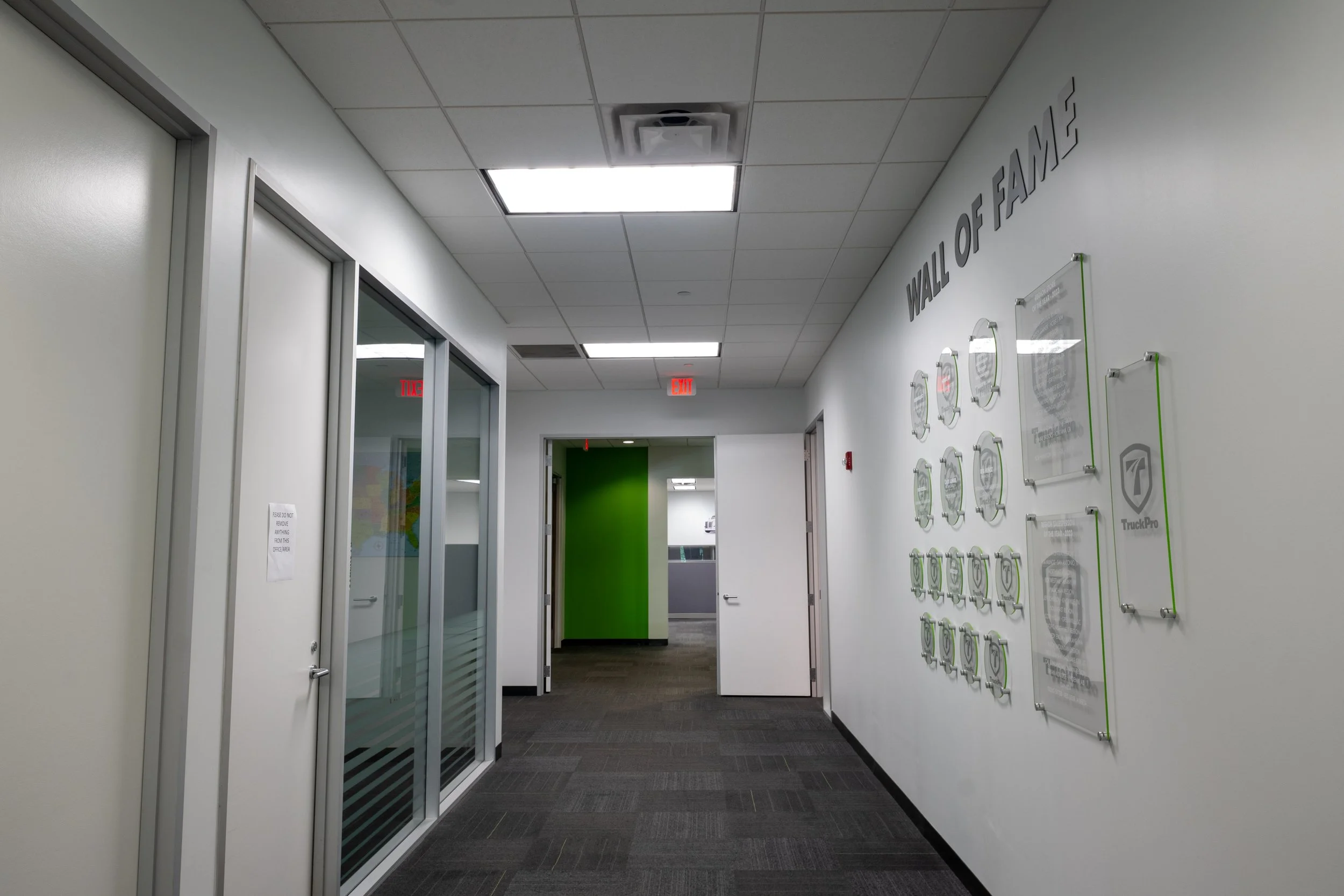 Office hallway with white walls, ceiling tiles, fluorescent lighting, a green door at the end, and wall-mounted display boards.