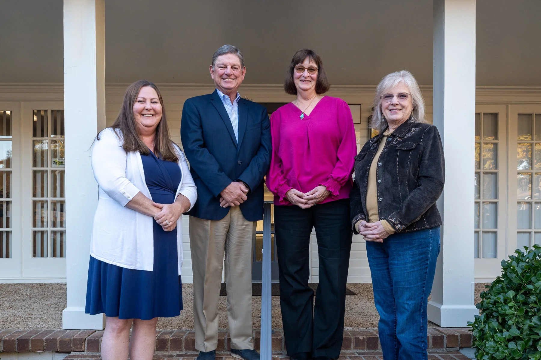 Four people standing in front of a white house, smiling, during daylight, dressed in business casual attire.