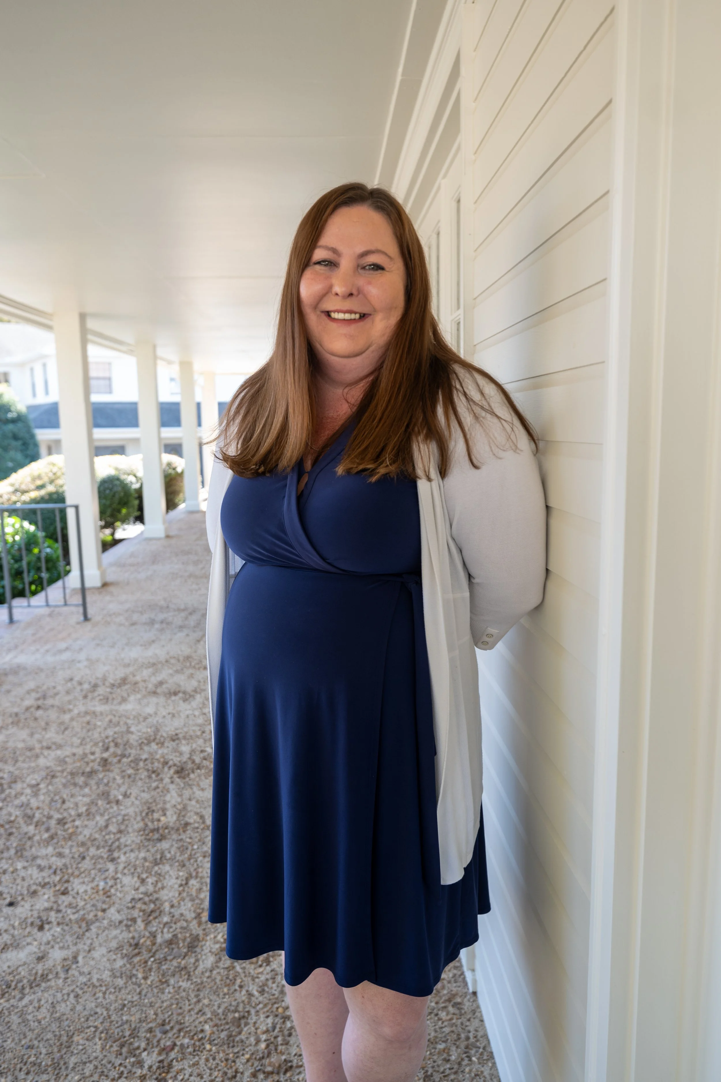 A smiling woman with long brown hair wearing a royal blue dress and a light gray cardigan, standing on a porch with white siding and a manicured garden in the background.
