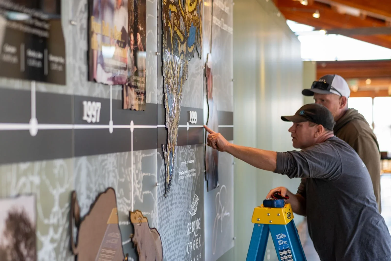 Two men are examining a large wall display with a world map. One man is pointing at a spot on the map while the other looks on. Both are wearing caps and casual outdoor clothing. One man is standing on a ladder.