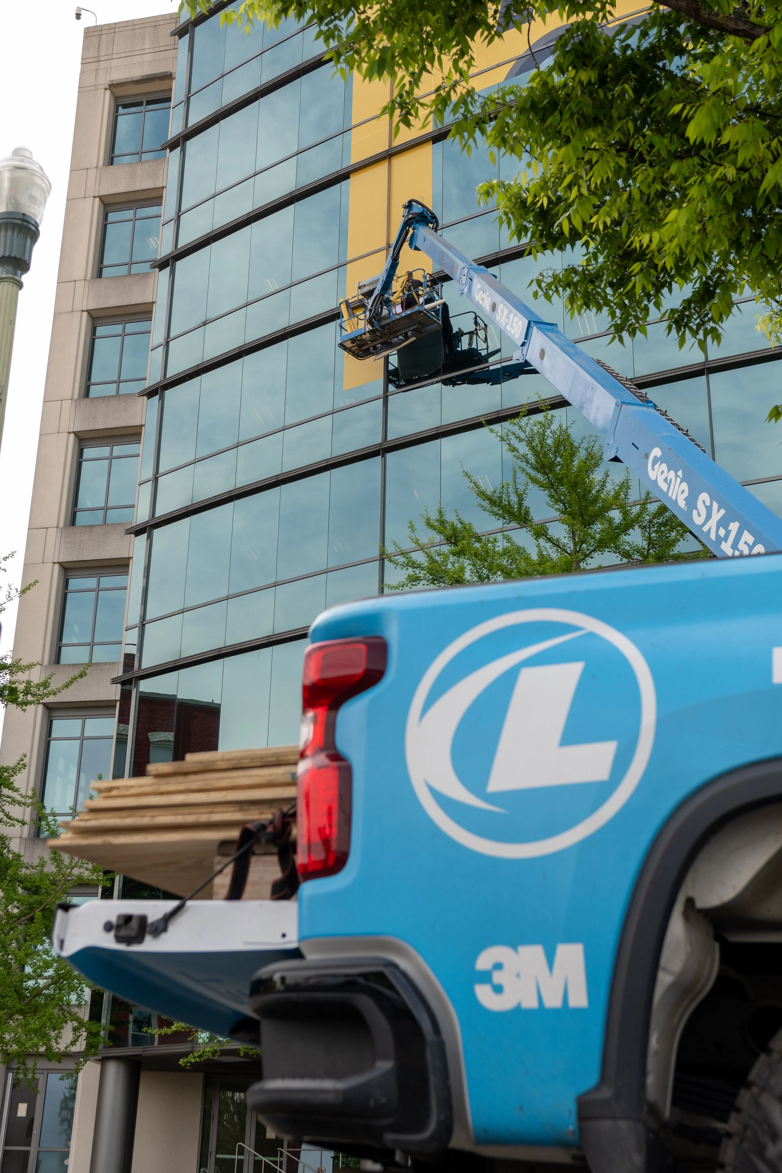 A construction worker is operating a blue boom lift at a building site. The lift is extended to work on the glass windows of a multi-story building, with green trees and a streetlamp visible nearby.