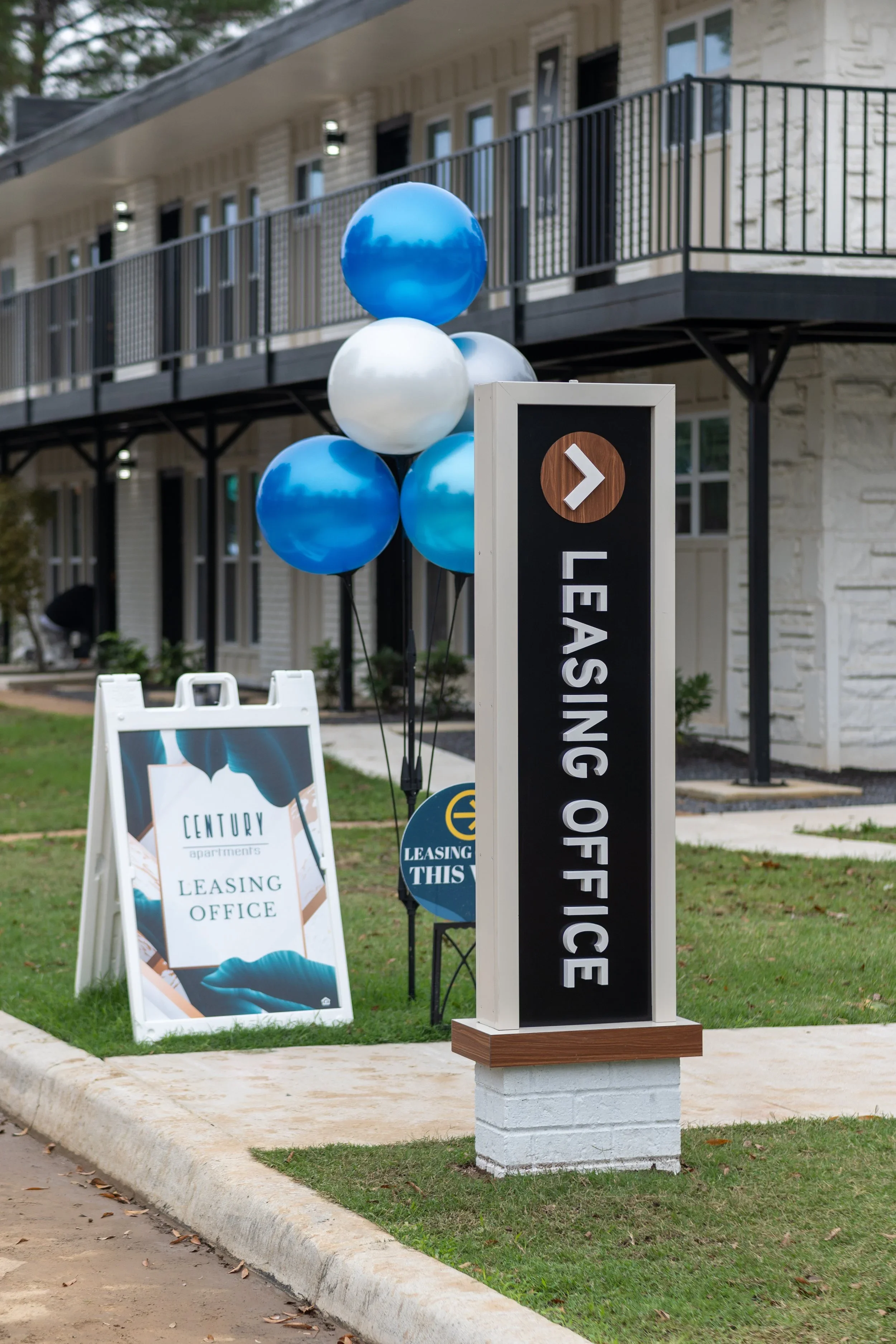 A sign and several balloons outside a building with residential stairs. The sign indicates leasing office for Century Apartments.
