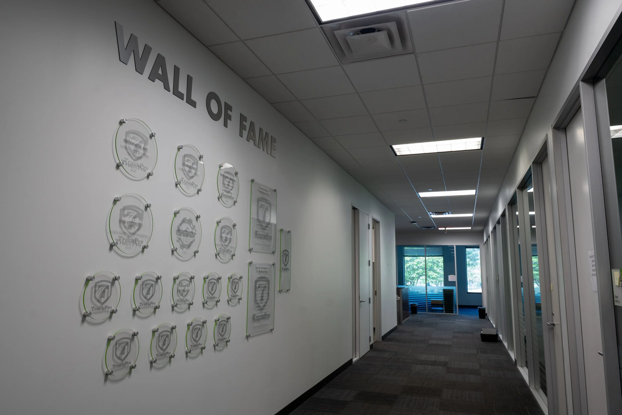 Indoor hall with wall display of plaques and awards, part of Wall of Fame, bright natural light from windows, ceiling lights, doors along the hall, modern office setting.