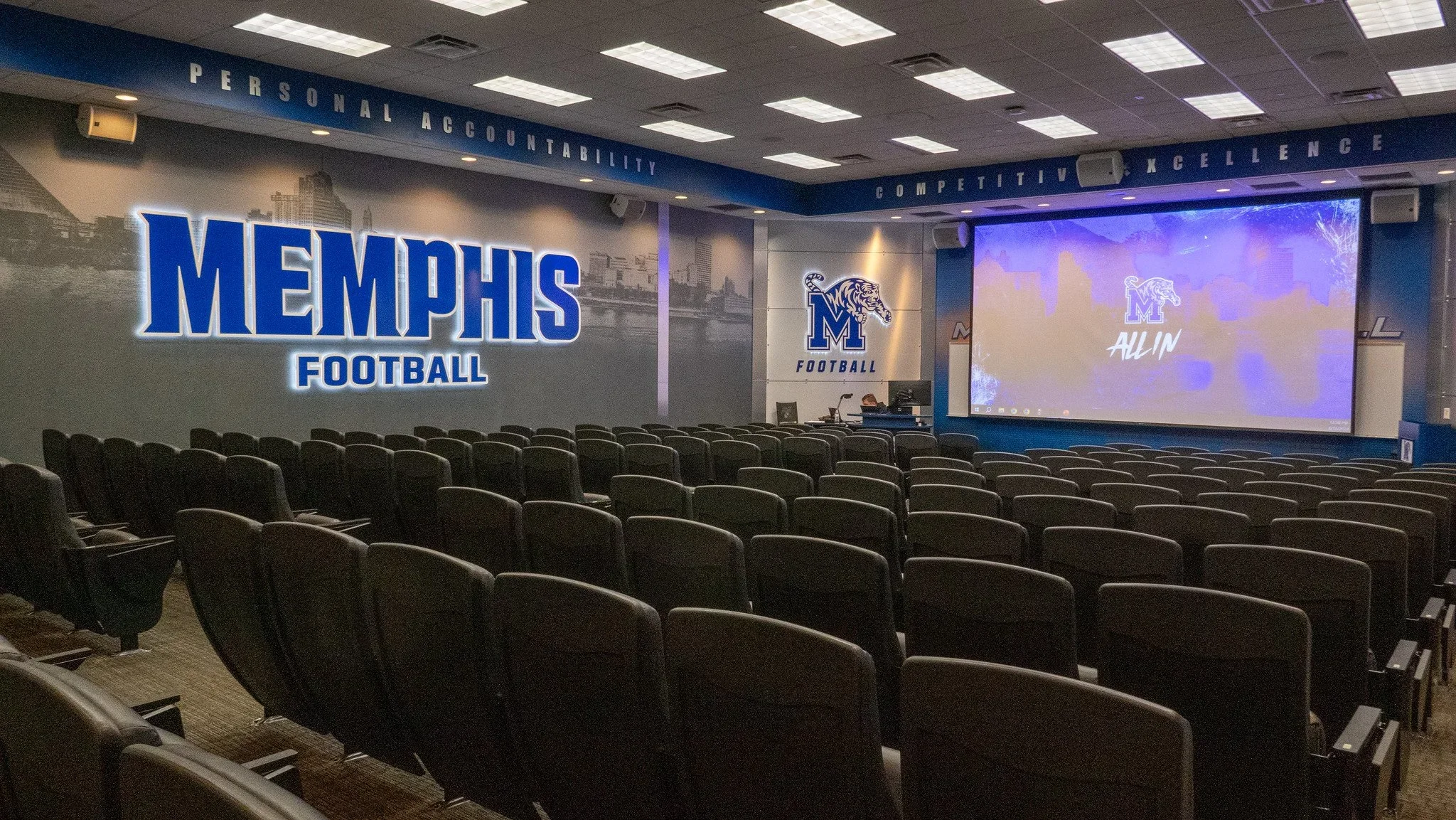 A university basketball classroom with chairs facing a large screen and wall displaying 'Memphis Football' and the Memphis Tigers logo, with motivational phrases like 'Personal Accountability,' 'Competitive,' and 'Excellence' on a banner overhead.
