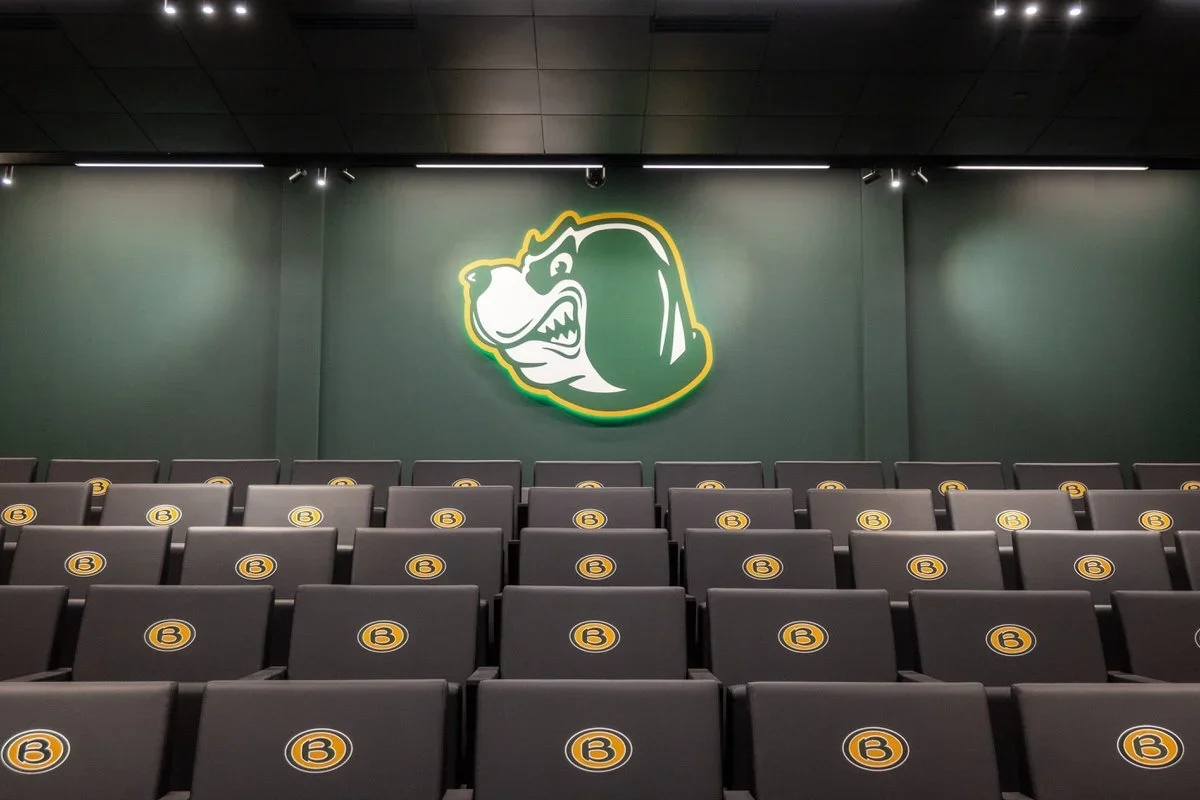 A room with rows of black seating chairs, each marked with a yellow circle and a letter B. On the wall at the front, there is a large illuminated logo of a bear's head in green, white, and yellow.