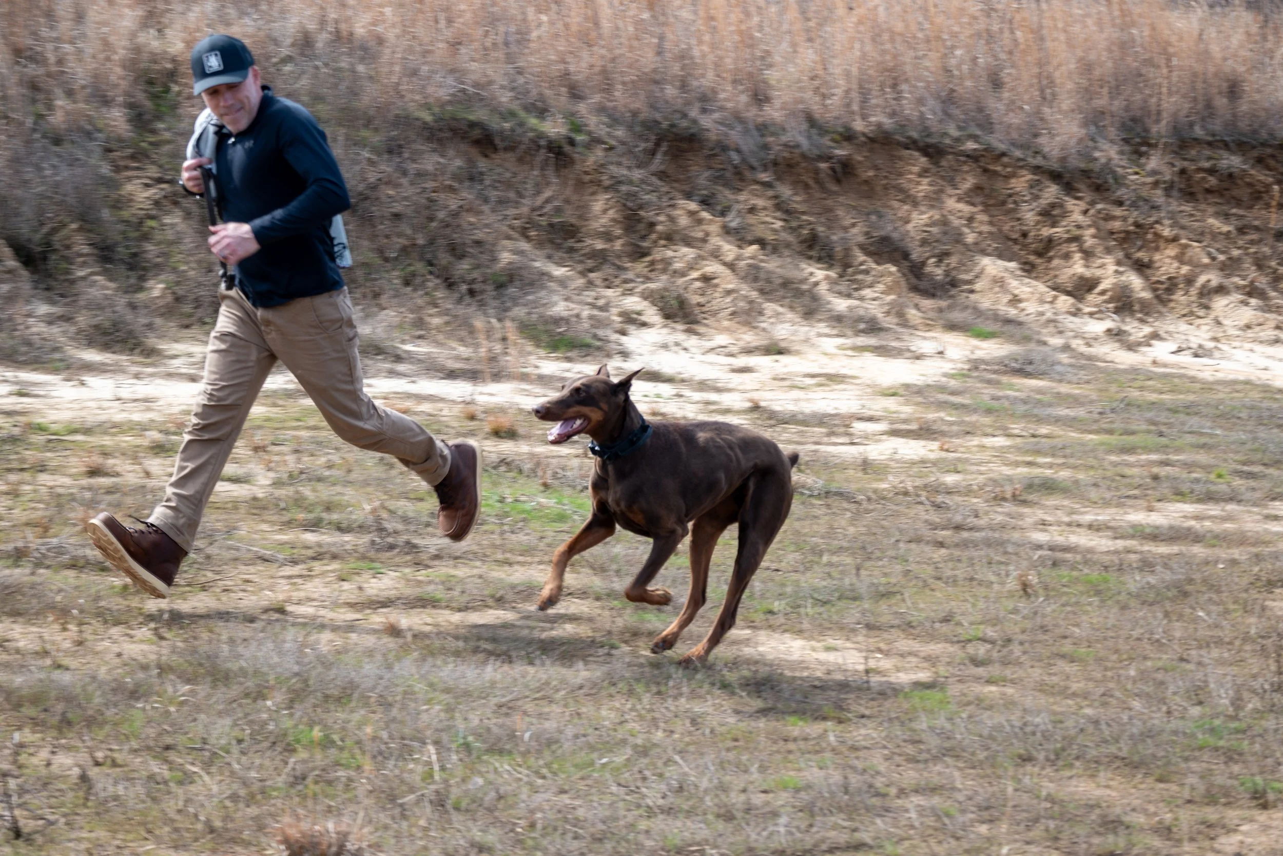 A man running outdoors with a brown dog, possibly a Doberman, during daytime in a grassy, partially wooded area with a hillside in the background.