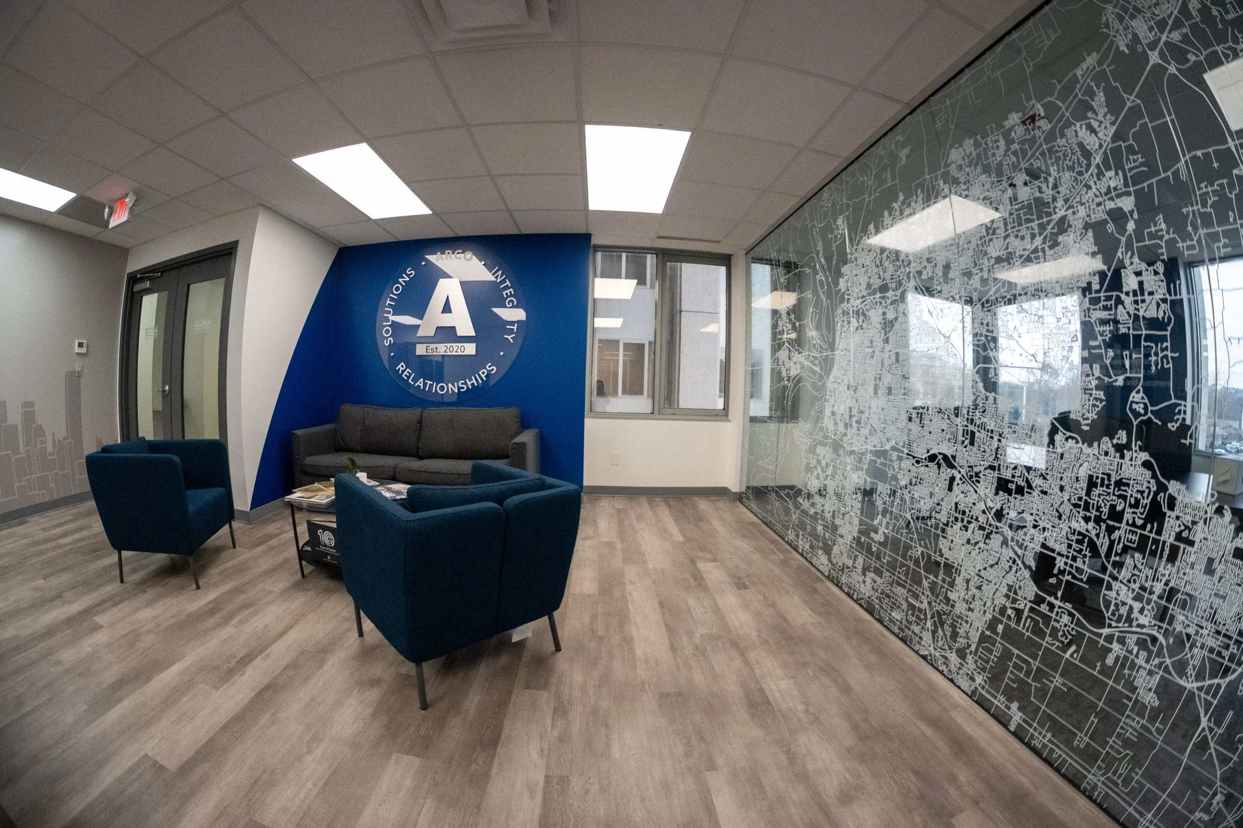An office waiting area with blue chairs and a gray couch arranged around a small table. A large wall sign with a white letter A and the words 'Solutions, Integrity, Relationships' on a blue background. Glass wall with a city map pattern, windows reve