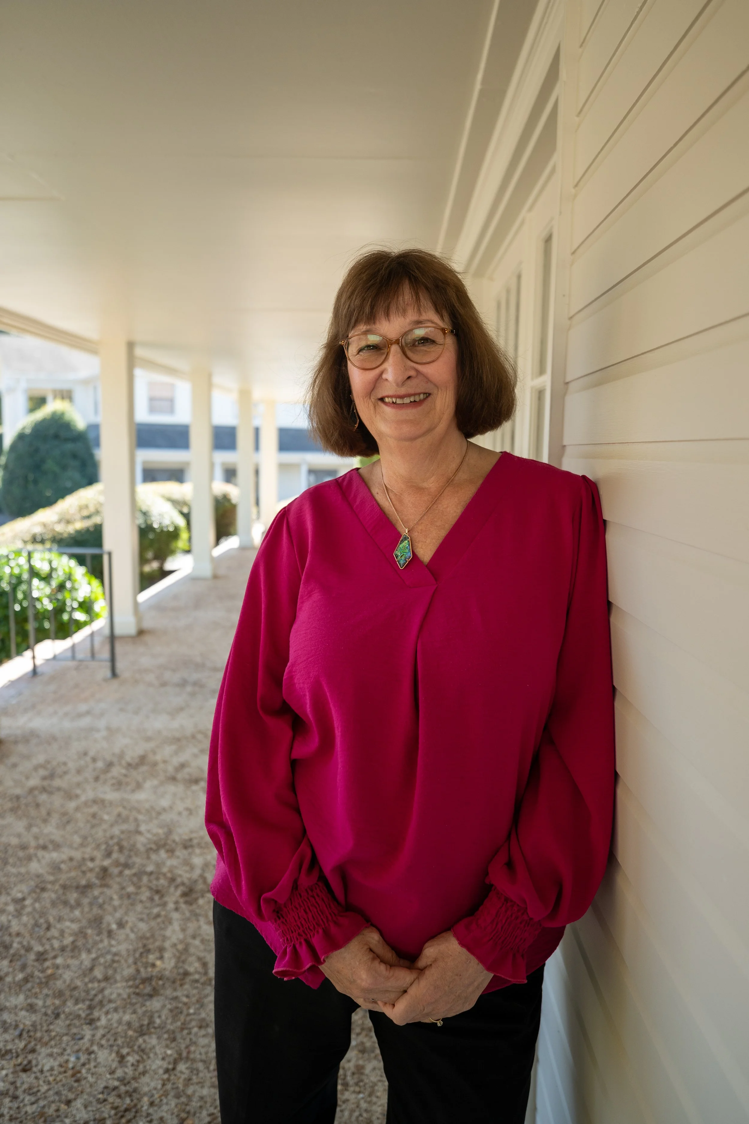 A smiling woman with glasses, wearing a pink blouse with ruffled sleeves, standing outside on a porch with her hands clasped. She has short brown hair and is wearing a necklace with a green gemstone. The porch has a beige floor, white support columns