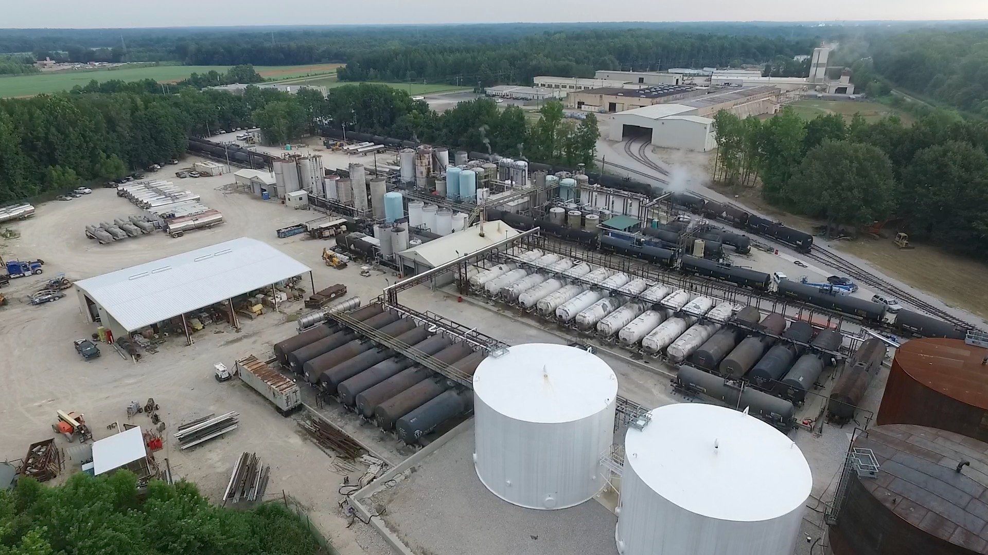 An industrial facility with large storage tanks, pipelines, and train tracks, surrounded by trees and green fields in the background.