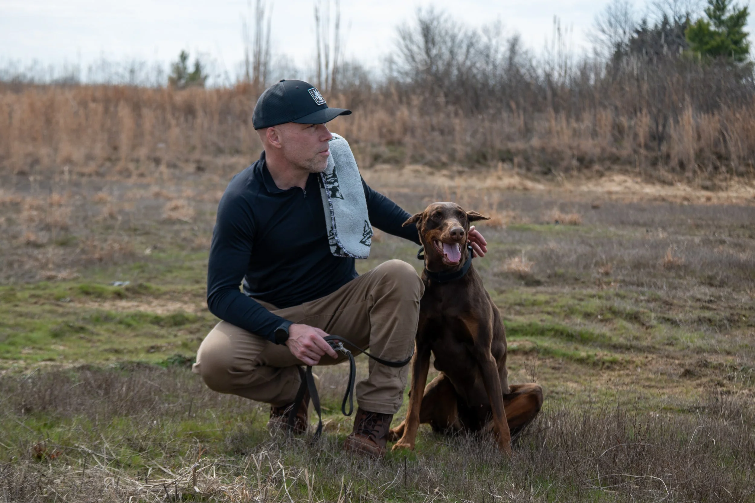 A man wearing a black cap and black long-sleeve shirt sitting on the grass outdoors with a brown dog sitting next to him, in a natural setting with dry grass and sparse trees in the background.