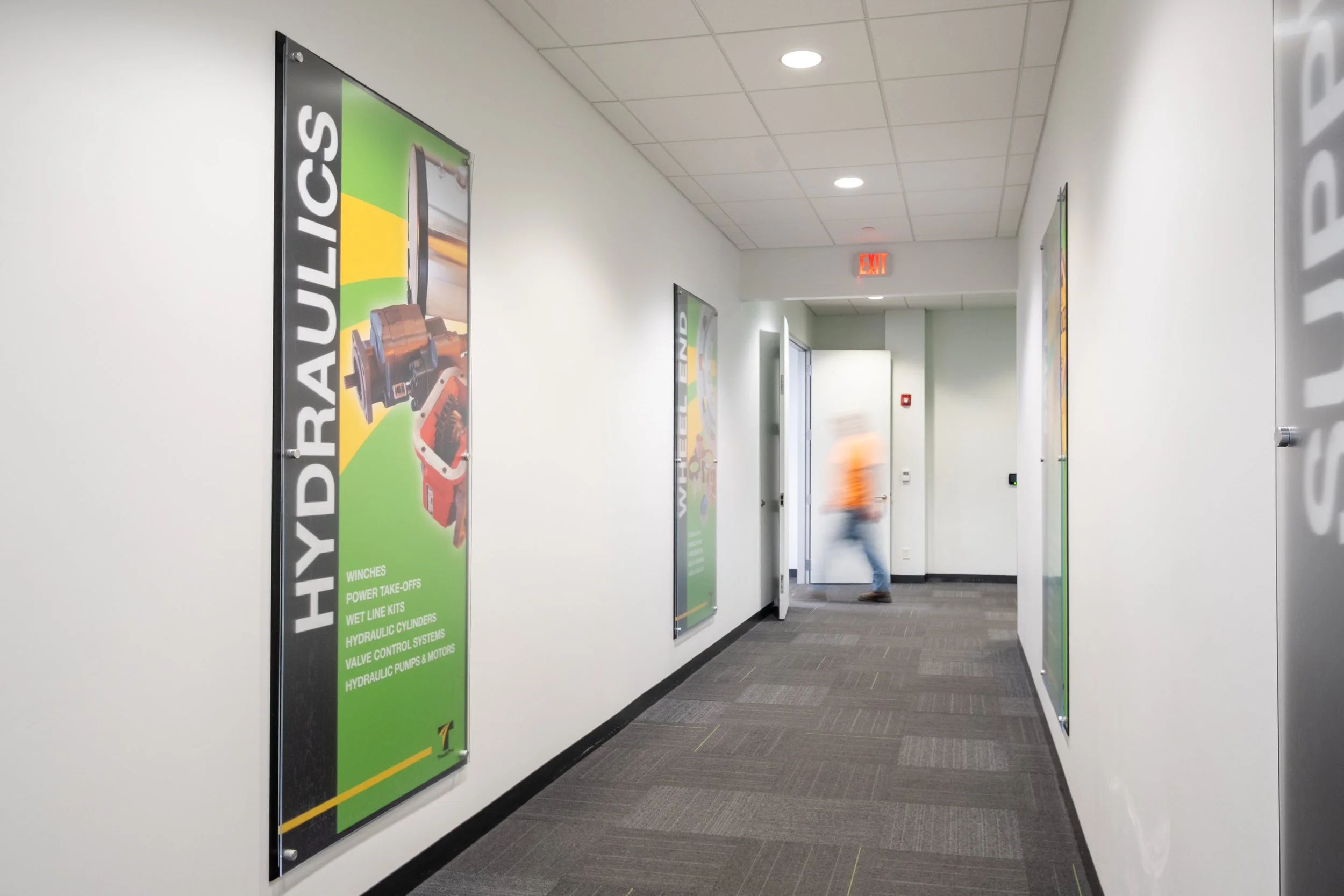 A hallway in a commercial building with white walls, a gray carpeted floor, and a white ceiling with fluorescent lights. On the left wall, there are multiple green posters advertising hydraulic parts and services. At the end of the hallway, there is 