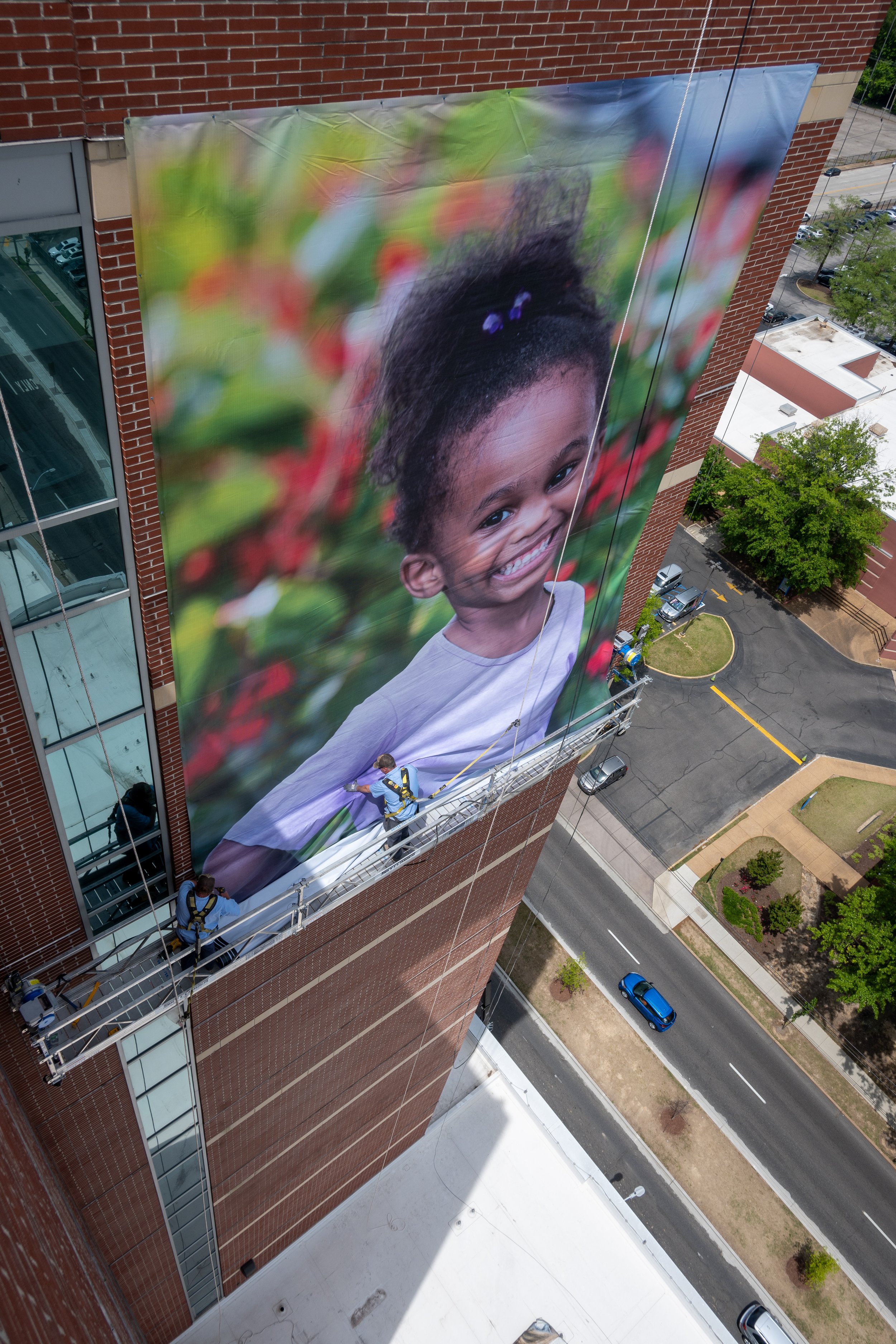 Workers installing a large outdoor banner with a smiling child's face on the side of a tall brick building.