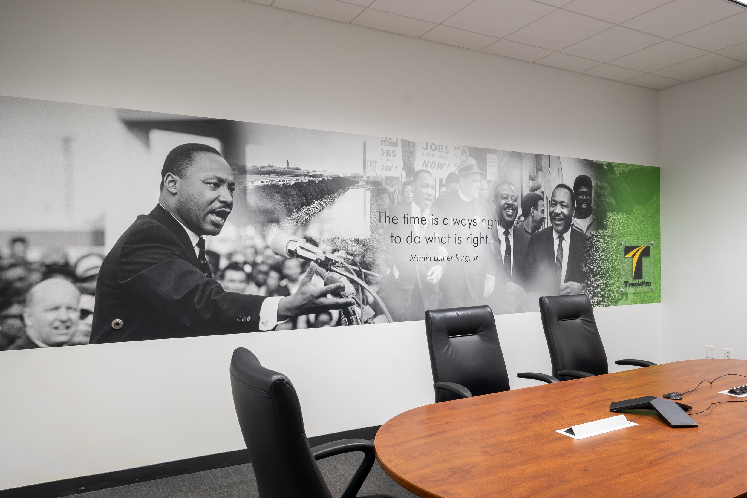 A conference room with a large wall mural featuring a black-and-white photo of Martin Luther King Jr. speaking at a rally, with a quote attributed to him. The room has a wooden conference table, black chairs, and some office supplies on the table.