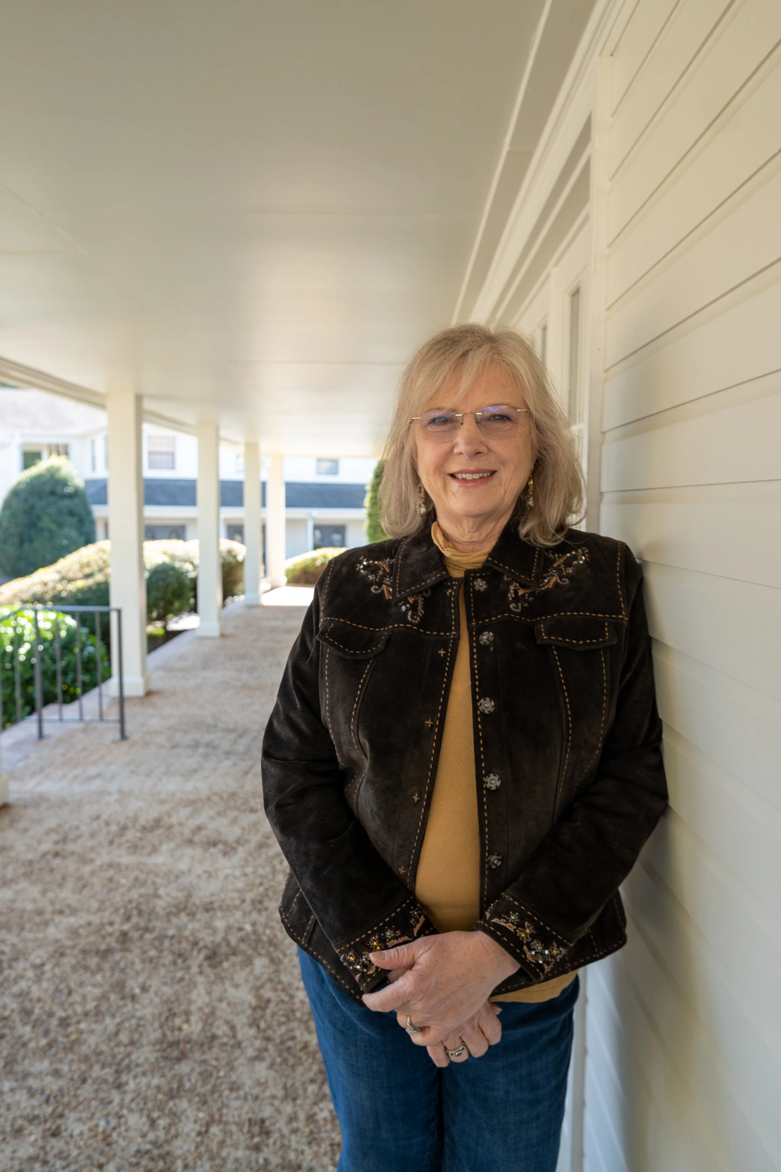 An elderly woman with gray hair and glasses smiling, standing next to a white house wall with siding, under a covered porch with columns, wearing a black embroidered jacket and blue jeans.