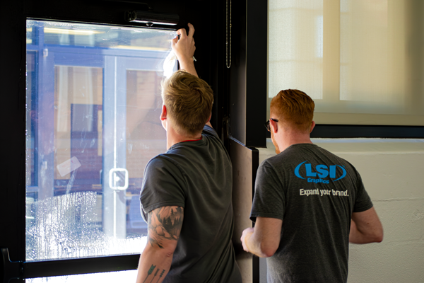 Two young men with short red hair, one wearing glasses and a gray T-shirt, the other with a tattooed arm in a black T-shirt, look out a window in an indoor setting.