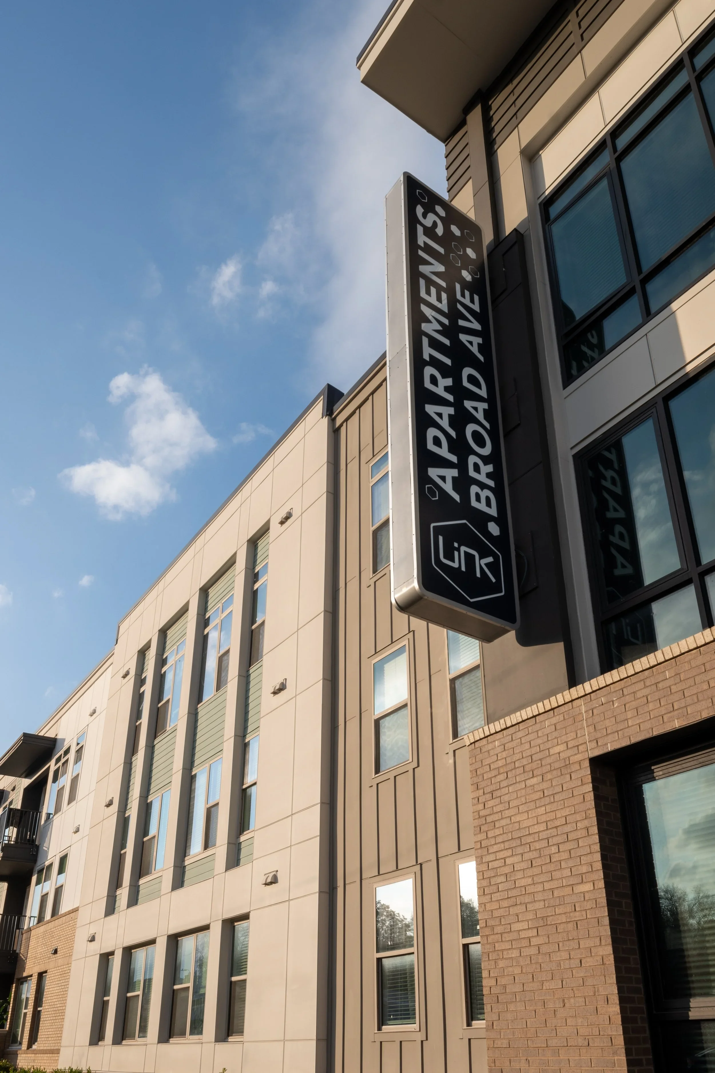A modern multi-story building with beige and brick exterior walls, large glass windows, and an overhead sign that reads "APARTMENTS - BROAD AVE." The sky is partly cloudy with some visible clouds.