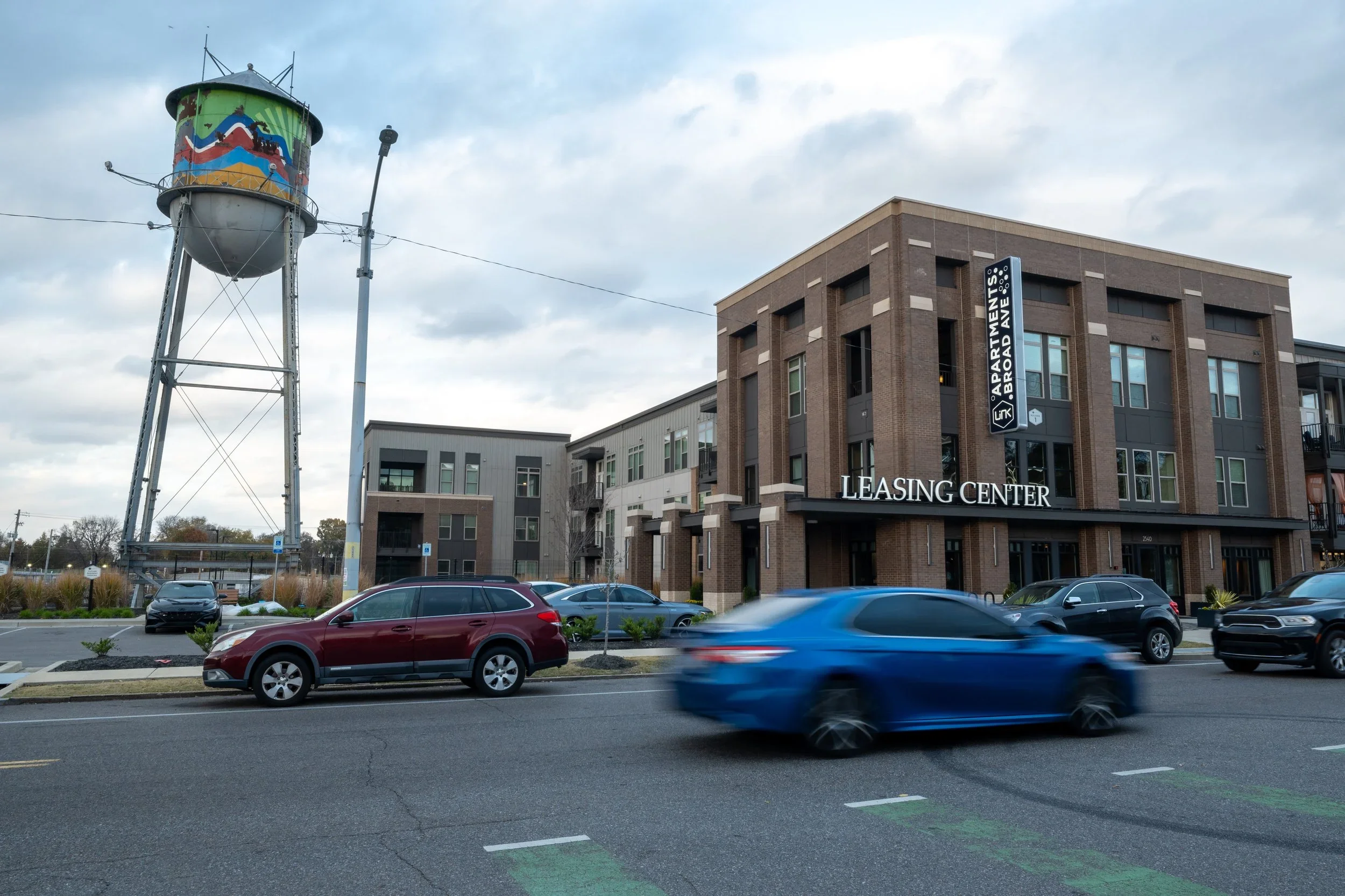 Parking lot with multiple cars in front of a brown brick building labeled 'Leasing Center' and 'Apartments on Broad'. A water tower with colorful graffiti art and a cloudy sky overhead.