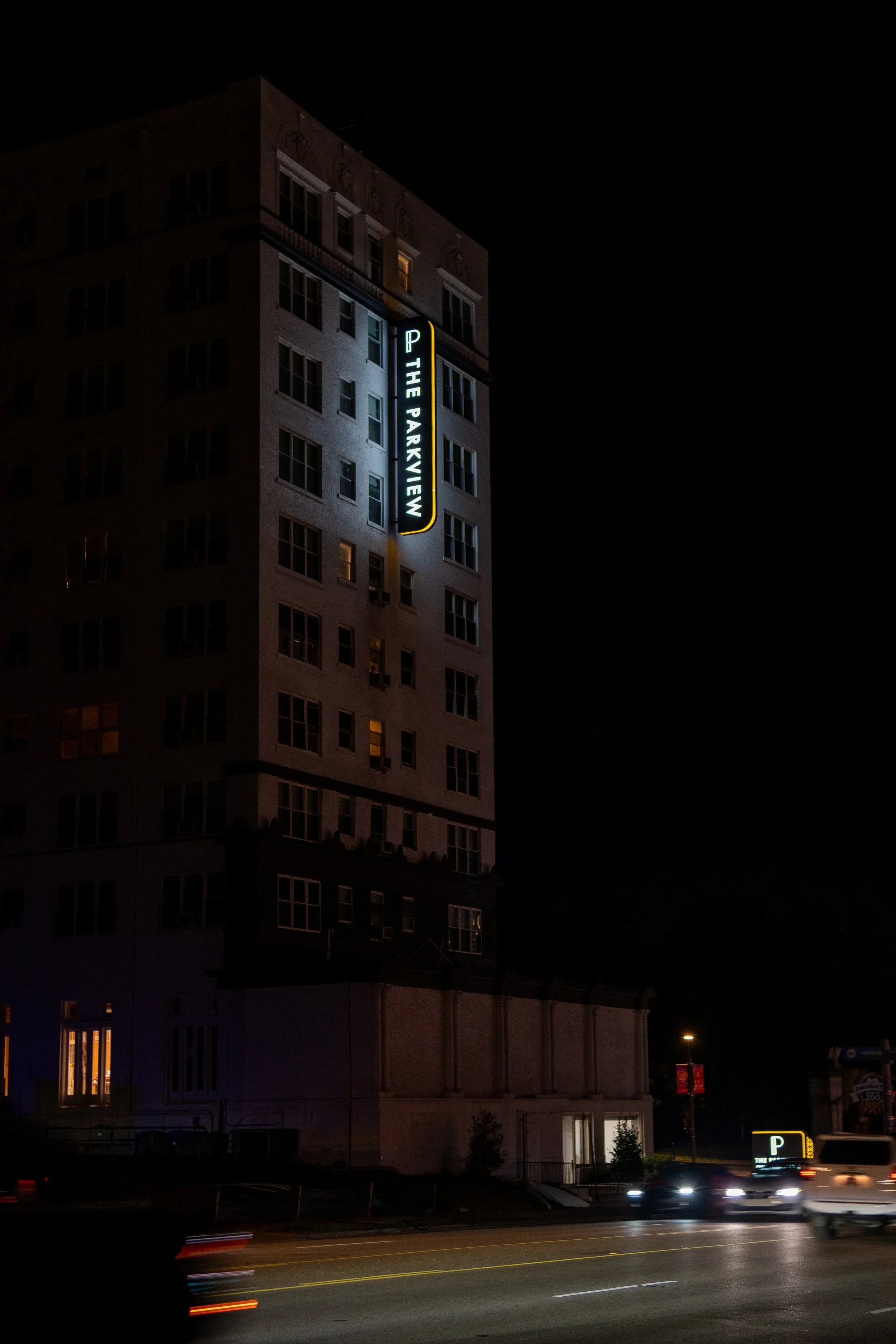 Nighttime city scene with a tall building displaying a vertical neon sign that reads 'THE PARKVIEW.' Cars are visible on the street with light trails from their movement, and a parking sign is visible in the foreground.