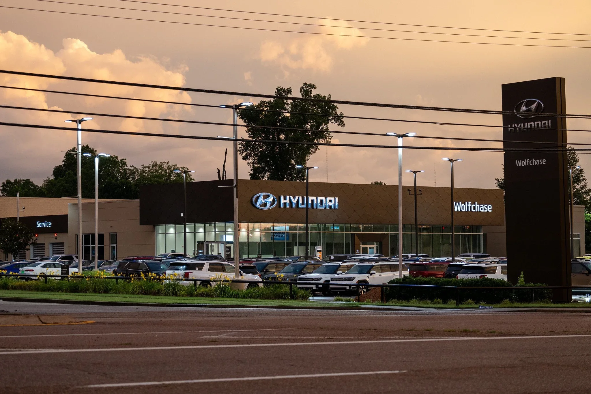 A Hyundai car dealership with illuminated signs and numerous parked cars in front during evening, with trees and cloudy sky in the background.