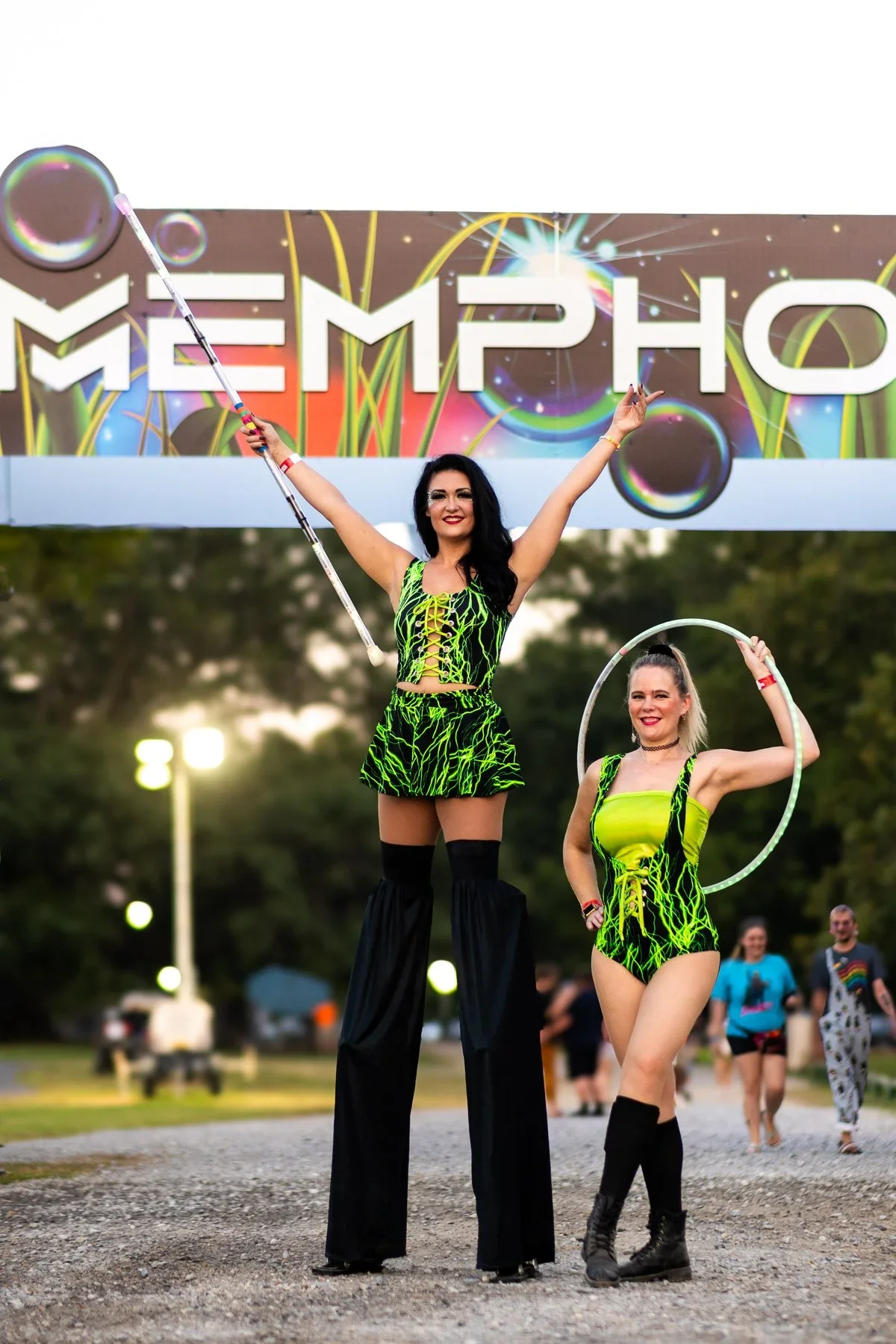 Two women dressed in neon green and black outfits with lightning patterns perform at a festival called Memphis. One is on stilts holding a pole, and the other holds a hula hoop. People in the background watch the performance.