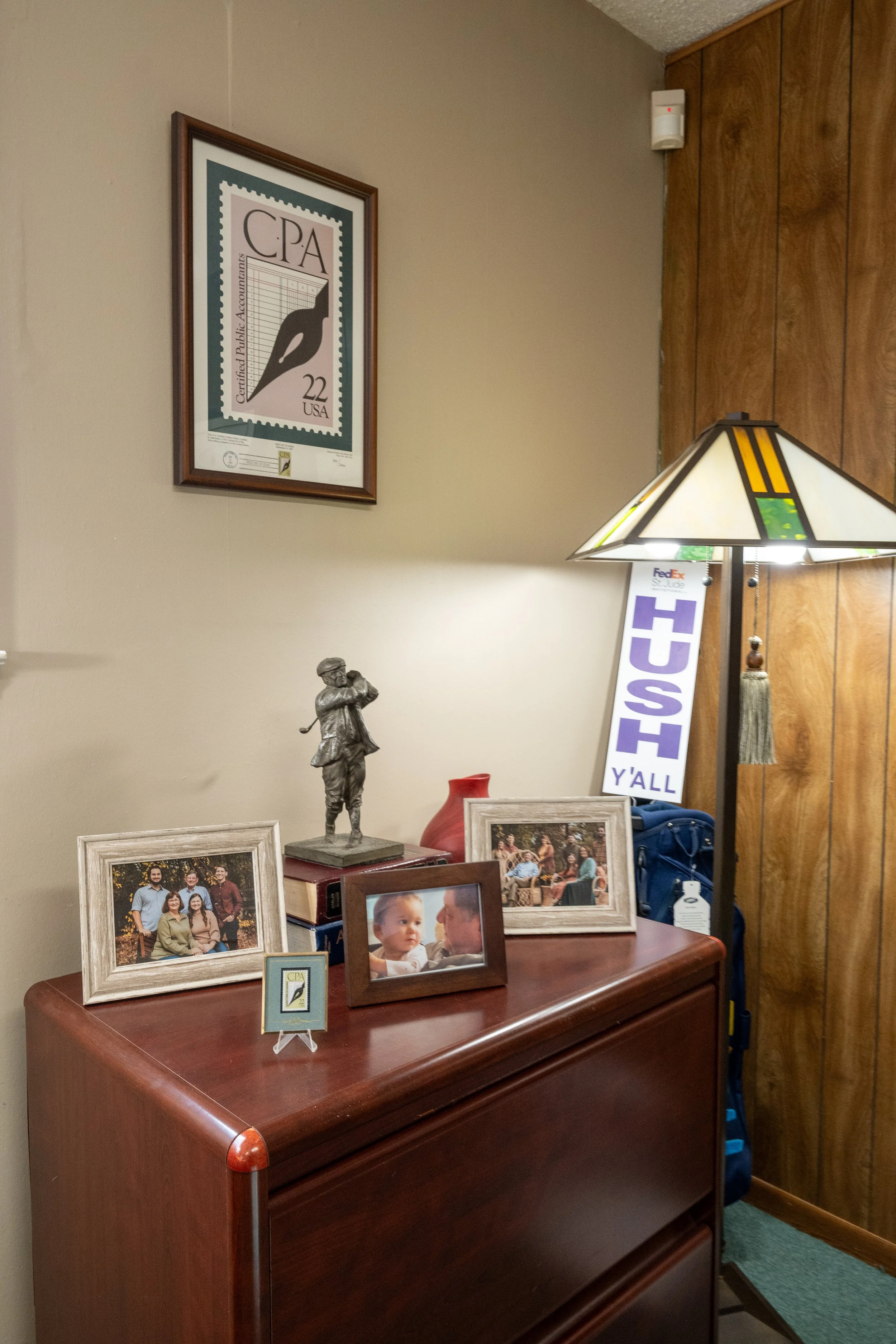 A wooden dresser with framed family photographs on it, a small statuette of a man holding a hat and pipe, a red vase, and small desk items. A lamp with a geometric stained glass design stands near the corner, along with a bag and a height measurement