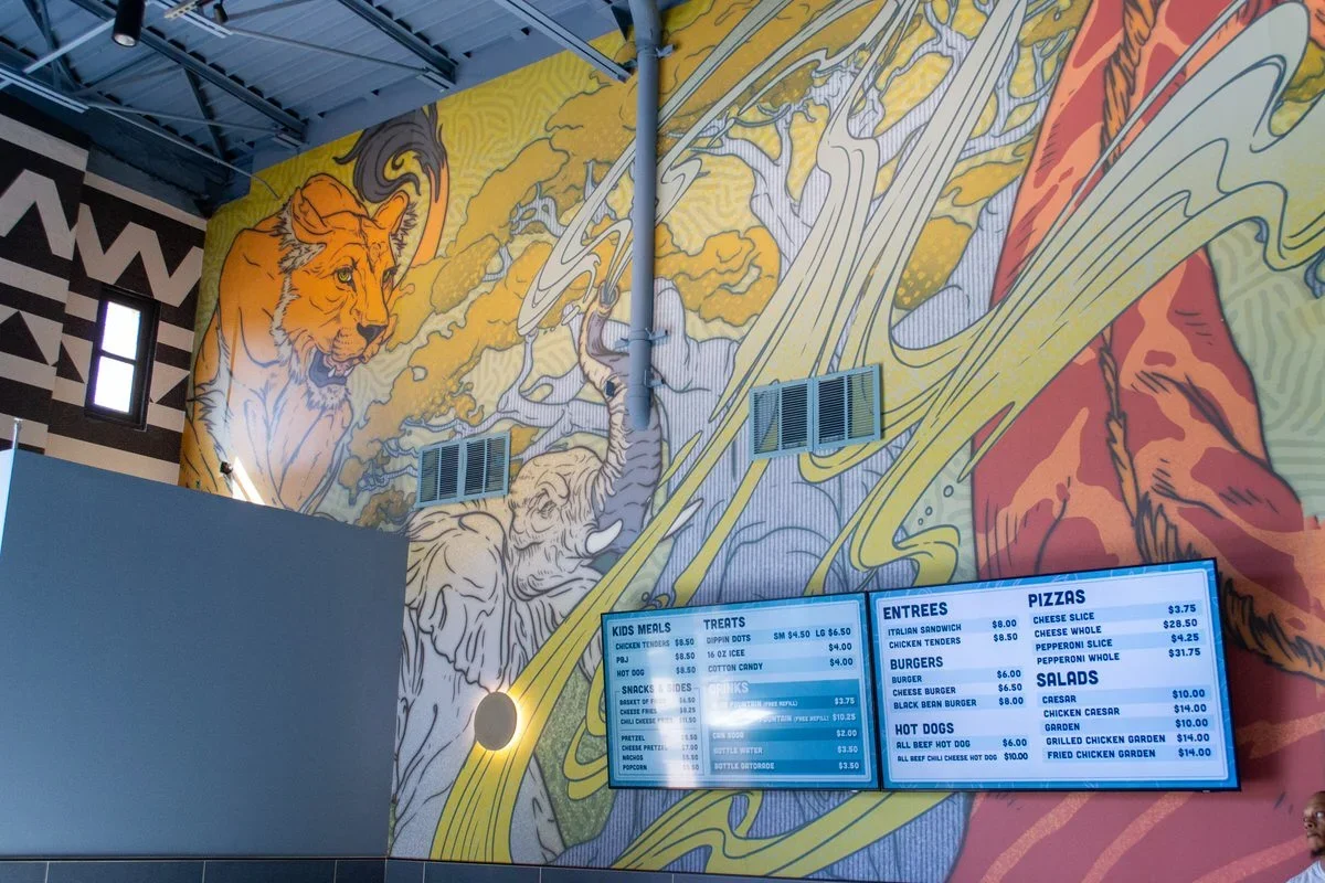 Colorful mural of a lion, elephant, and other animals on a wall inside a restaurant, with digital menu screens displaying food options and prices.