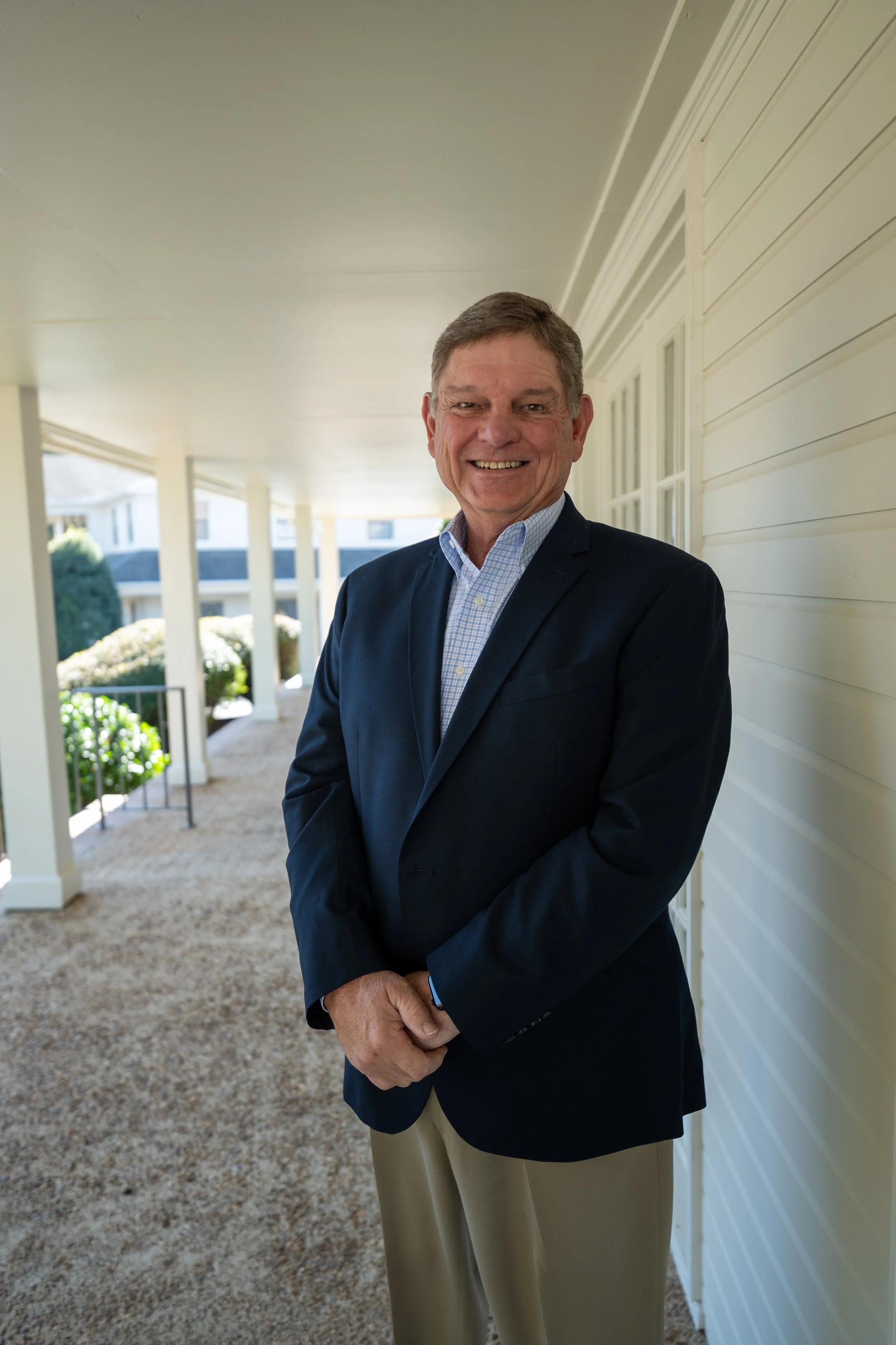 A smiling middle-aged man in a navy blazer, light blue checkered shirt, and khaki pants standing on a porch with white siding and windows. There are shrubs and a house in the background.