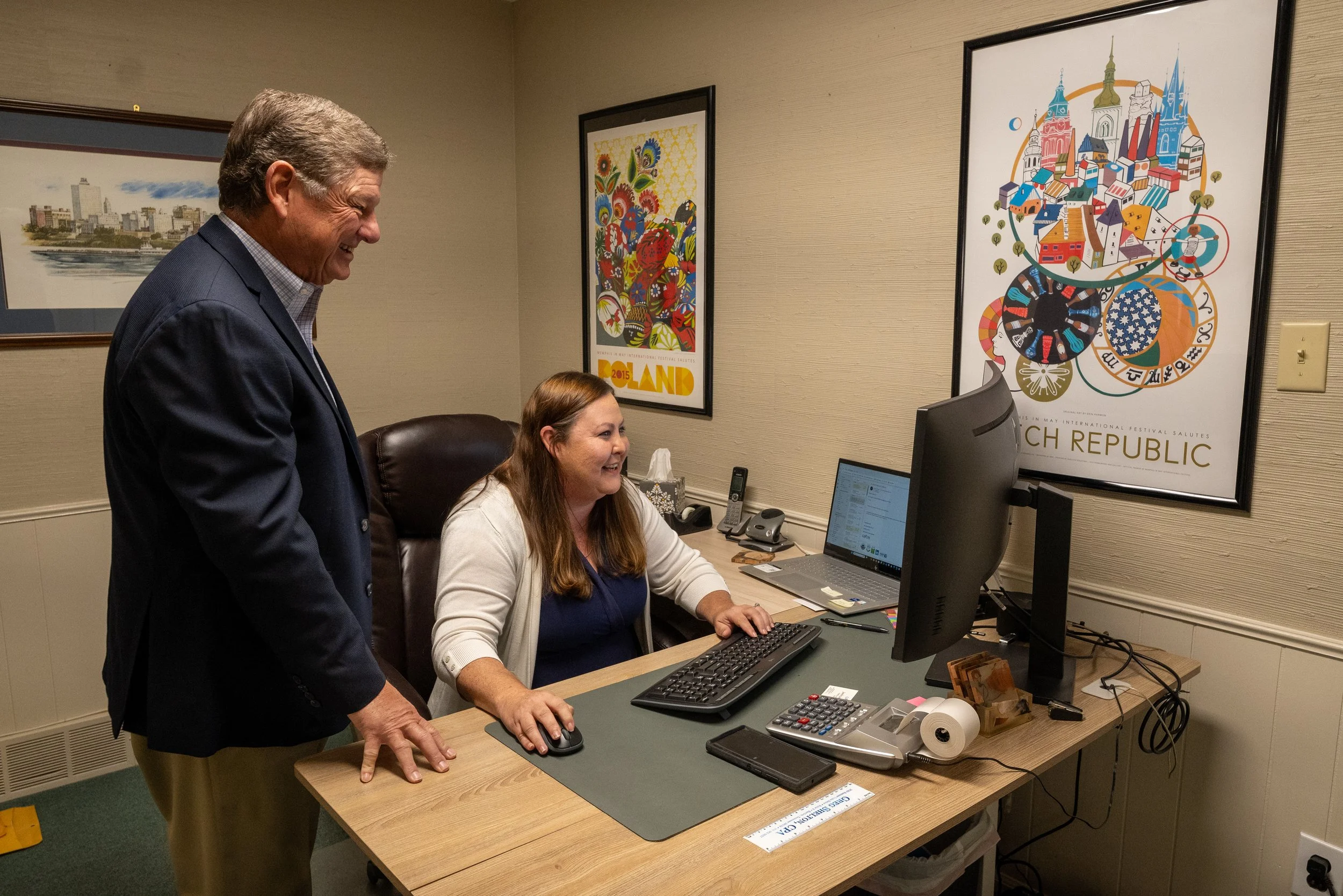 A man and a woman are in an office, looking at a computer screen. The man is standing and the woman is seated at a desk, typing on a keyboard. The wall has colorful posters, including one with a cityscape and another with abstract artwork. The desk h