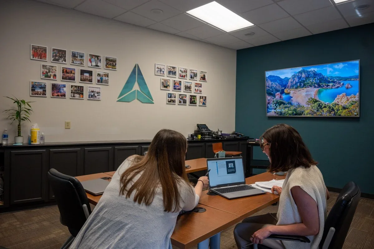 Two women seated at a conference table working on a laptop in a meeting room with picture frames on the wall and a large monitor displaying a scenic landscape.