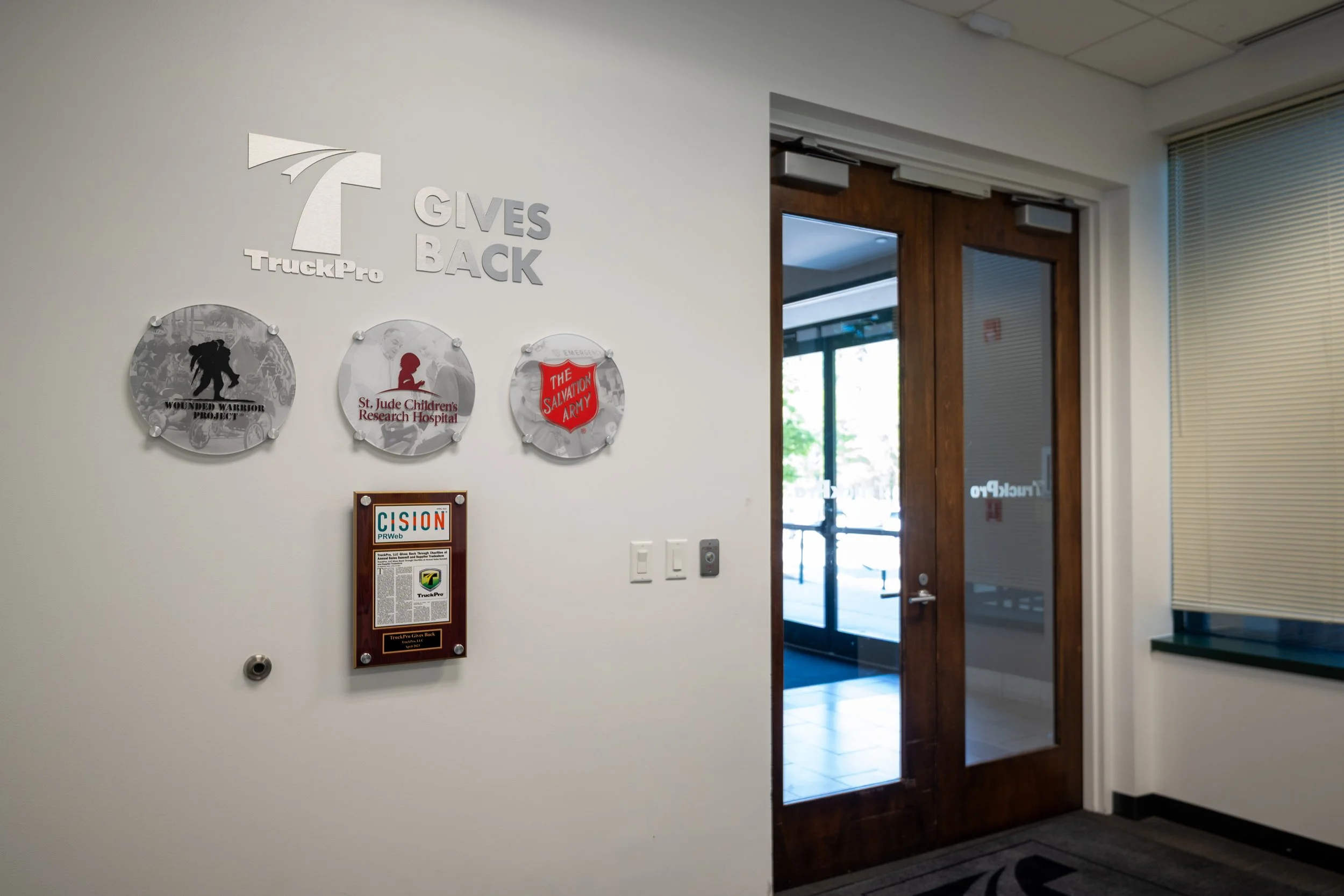 Indoor scene with a white wall displaying logos and plaques, near a wooden door with glass panel and a window with blinds.