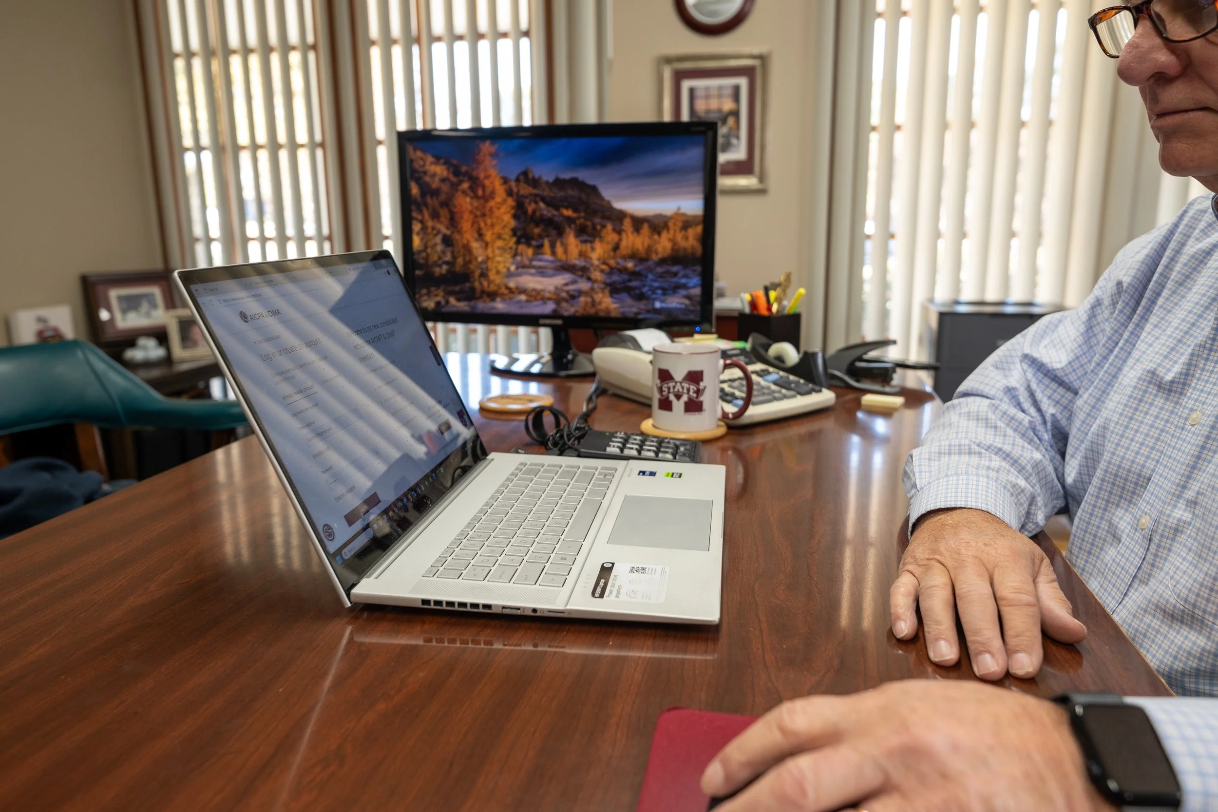 A man wearing glasses sitting at a wooden desk with a laptop, a computer monitor displaying a scenic landscape, a coffee mug with a college logo, and various office supplies, in a room with vertical blinds and framed pictures.