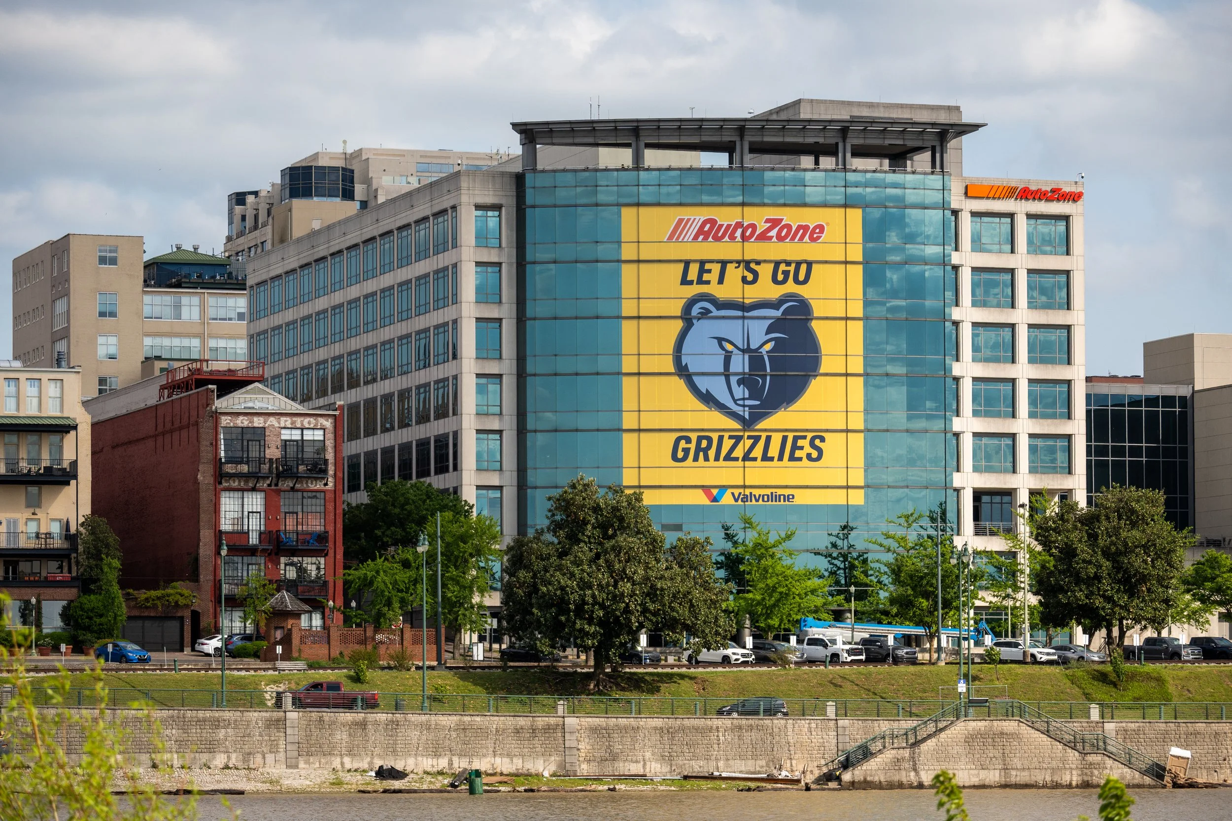 Cityscape with a large billboard on a building that features an advertisement for the Memphis Grizzlies basketball team. The billboard shows a grizzly bear face and the text "LET'S GO" and "GRIZZLIES." The city includes various mid-rise buildings, tr