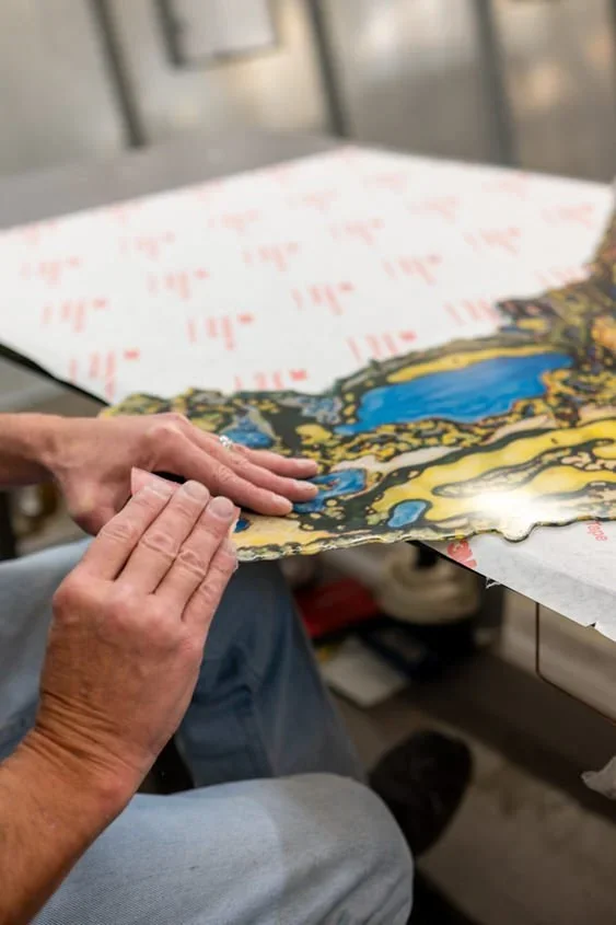 Close-up of a person's hands touching a glossy, colorful art paper with blue and yellow patterns, sitting at a table in a workshop.