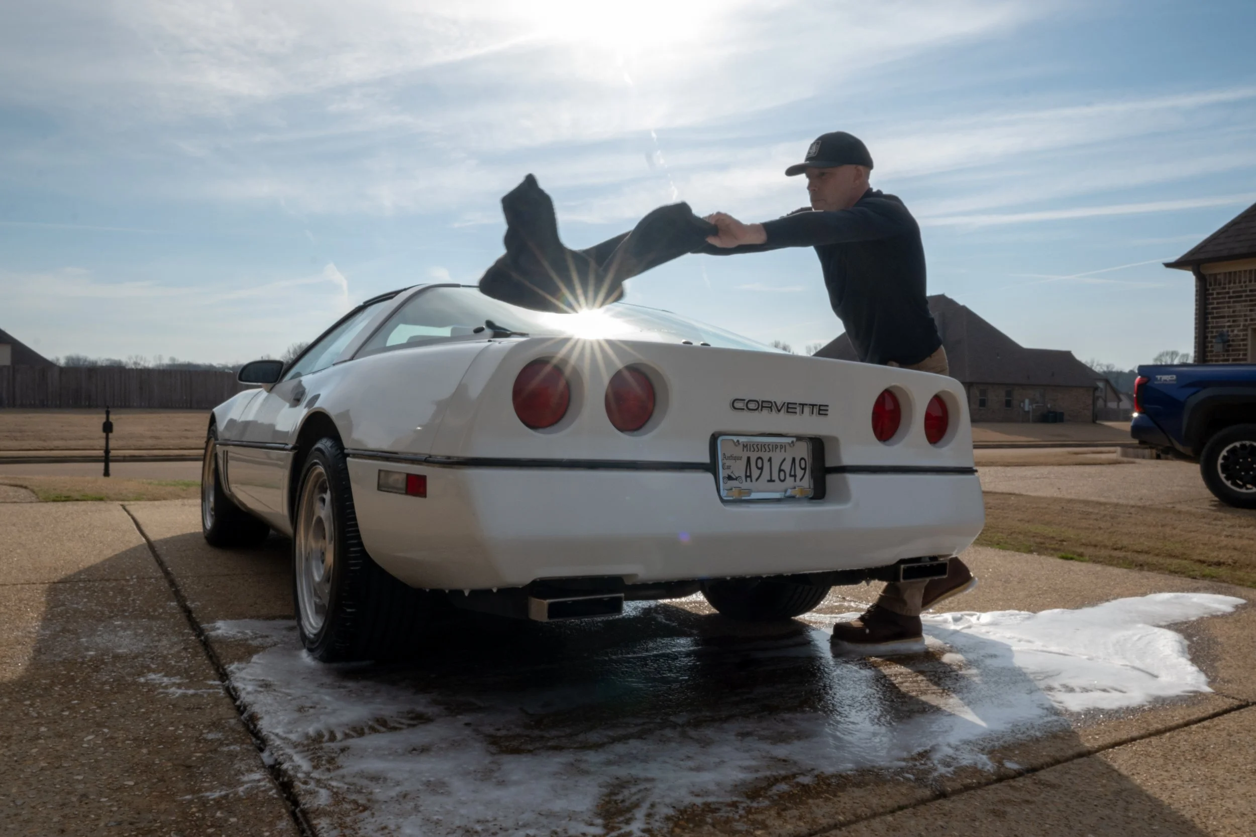 A man is washing a white Chevrolet Corvette sports car in a driveway. The man is removing a black cloth from the car's front. The sky is partly cloudy, and puddles of water are on the ground.