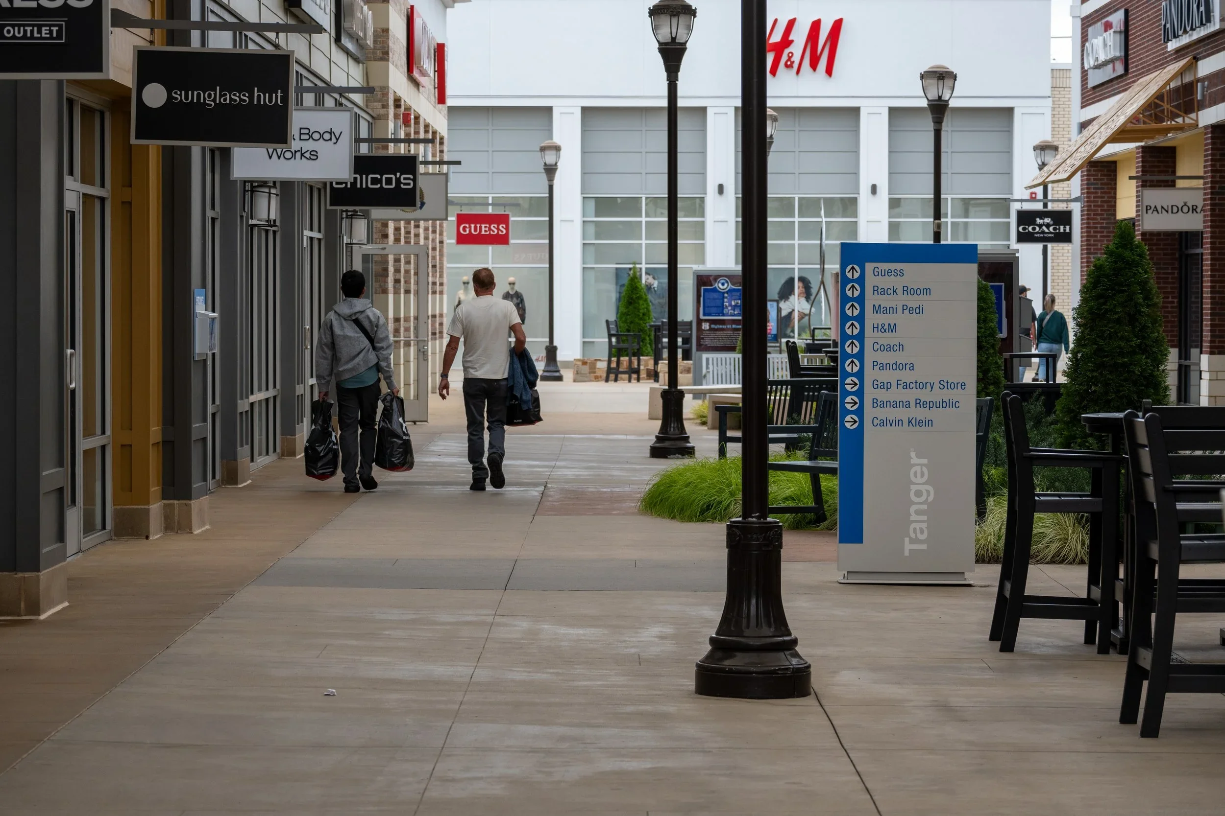 A shopping mall corridor with several storefront signs, including Sunglass Hut, Chico's, Guess, Coach, Pandora, and H&M. There are people walking and sitting, and a directional sign listing various stores and brands.