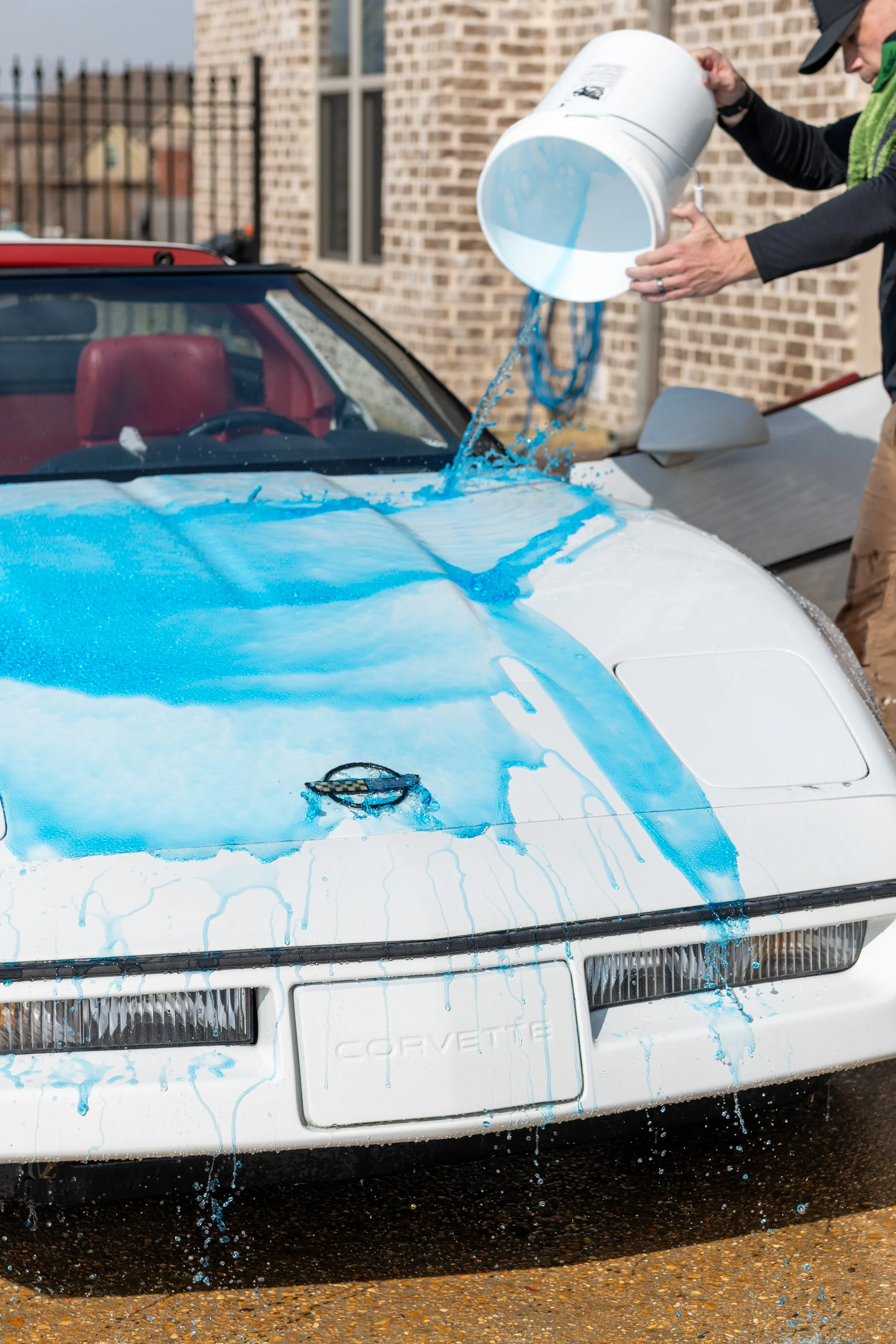 Person pouring blue liquid onto white vintage Corvette car with emission of drips and splashes.