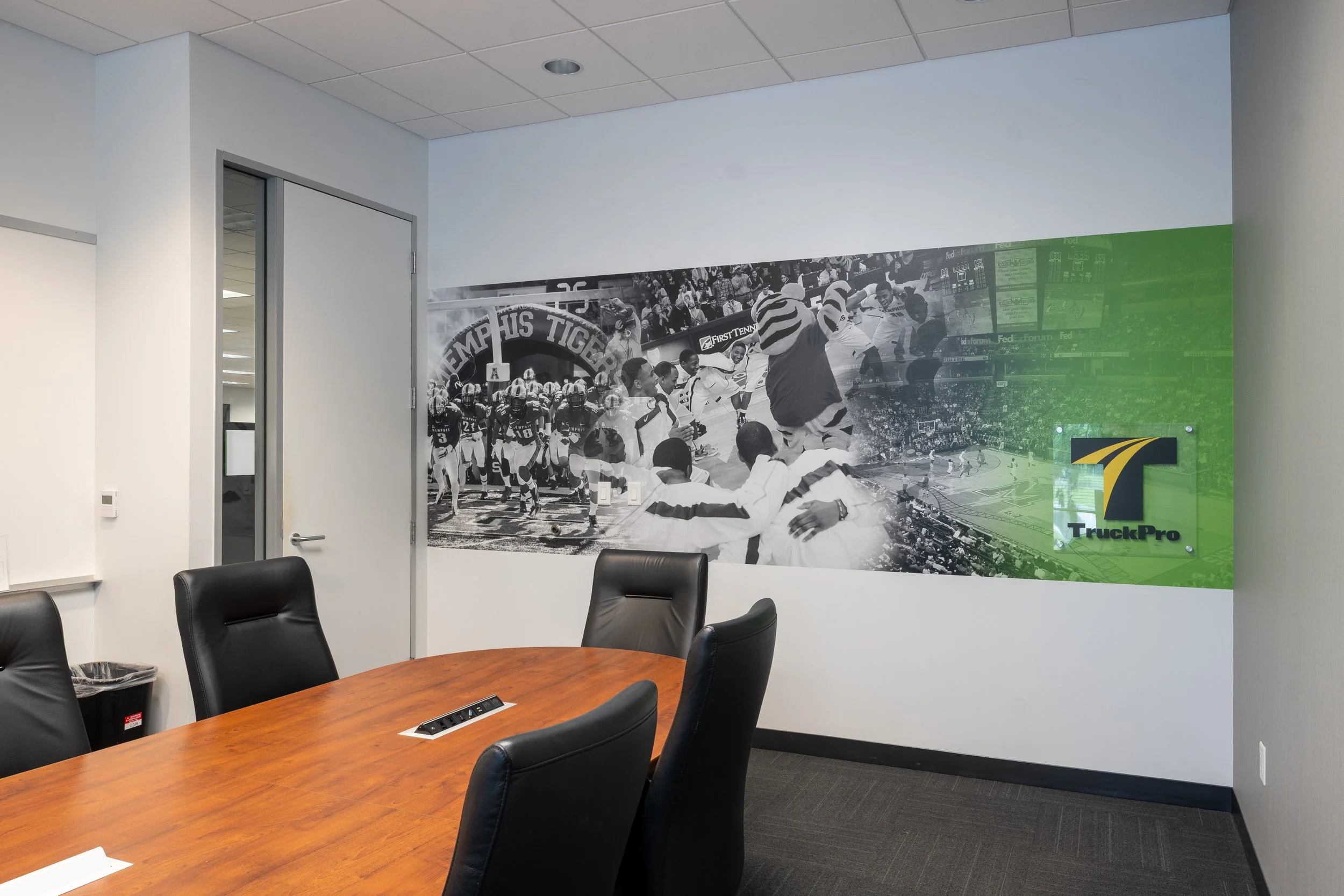 Conference room with a wood table and black chairs, featuring a large mural of a black-and-white sports photograph on the wall. The mural depicts a football team and a mascot, with a green accent on the right side, along with the logo and name 'Truck