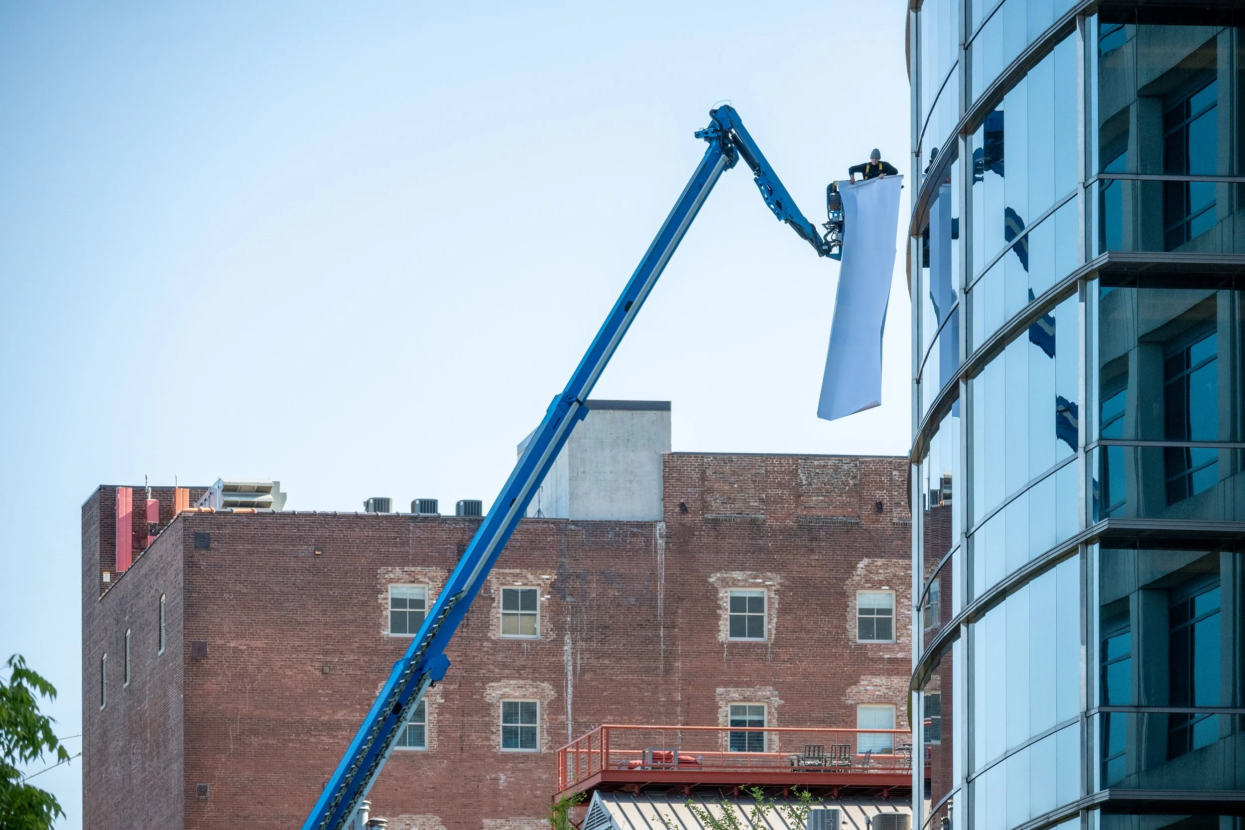 A worker on a hydraulic lift installing a large white banner on the exterior of a modern glass office building, with older brick buildings visible in the background.