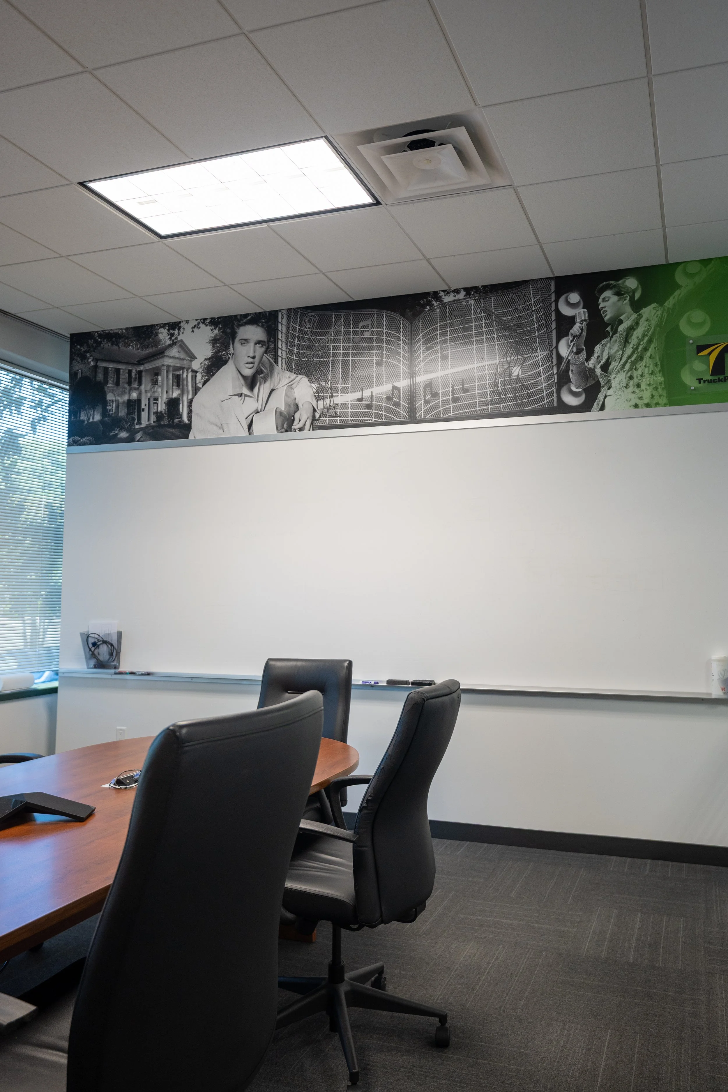 Office conference room with a wooden table and black leather chairs. A whiteboard is mounted on the wall, and a large black-and-white mural features Elvis Presley and a historic building. The ceiling has a rectangular light panel and an air vent, and