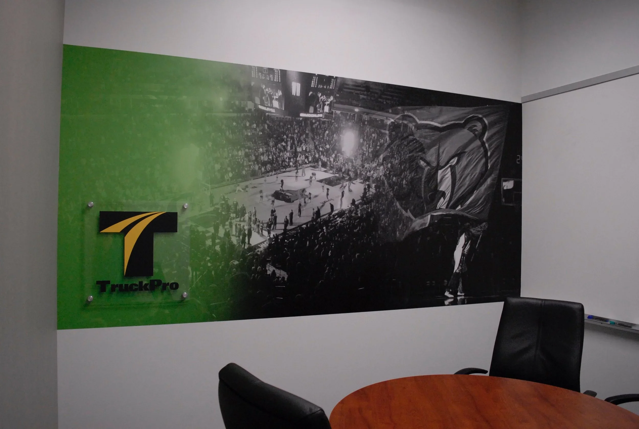 A conference room with chairs around a wooden table and a large poster on the wall depicting a sports arena filled with spectators, advertising a TruckPro event.