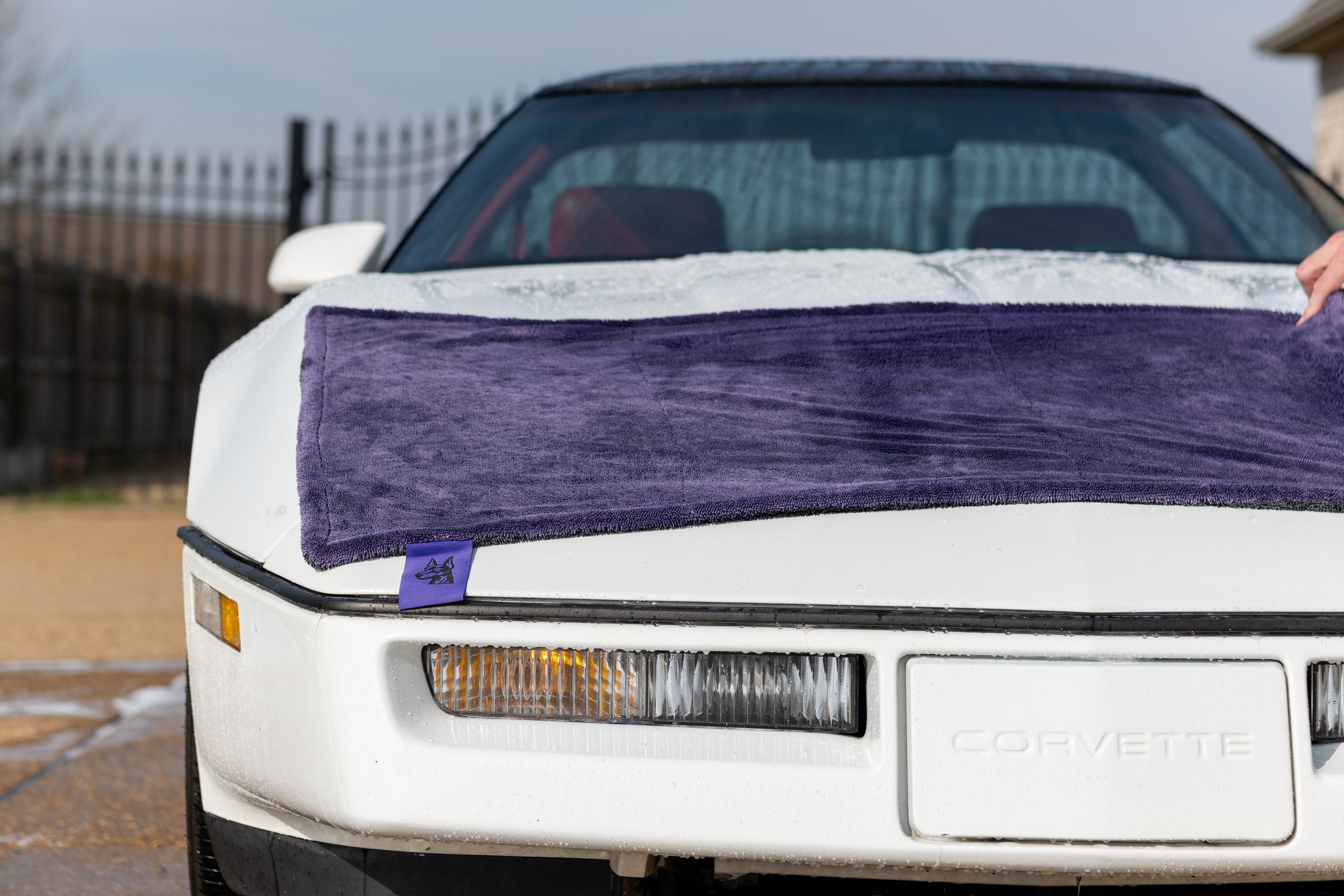 Front of a white Chevrolet Corvette car with a purple towel draped over the hood, wet from water, outside during the day.